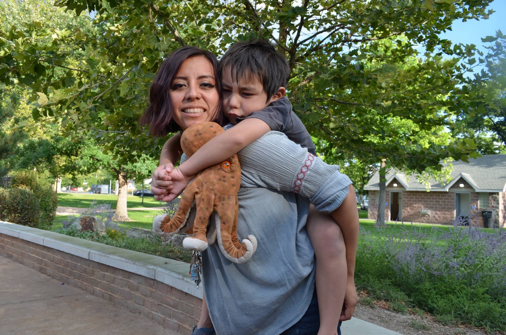 Woman outdoors with child holding teddy bear on her back, she is smiling