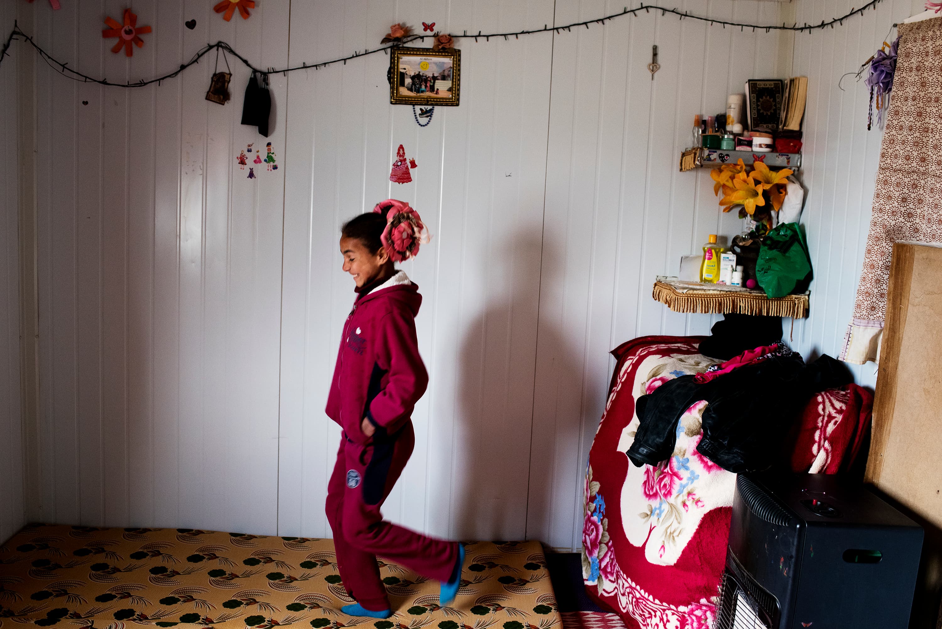 Solana, 11, walks through one of the rooms inside the family's Zaatari shelter. Behind her, a portrait of the whole family hangs among lights and decorations.