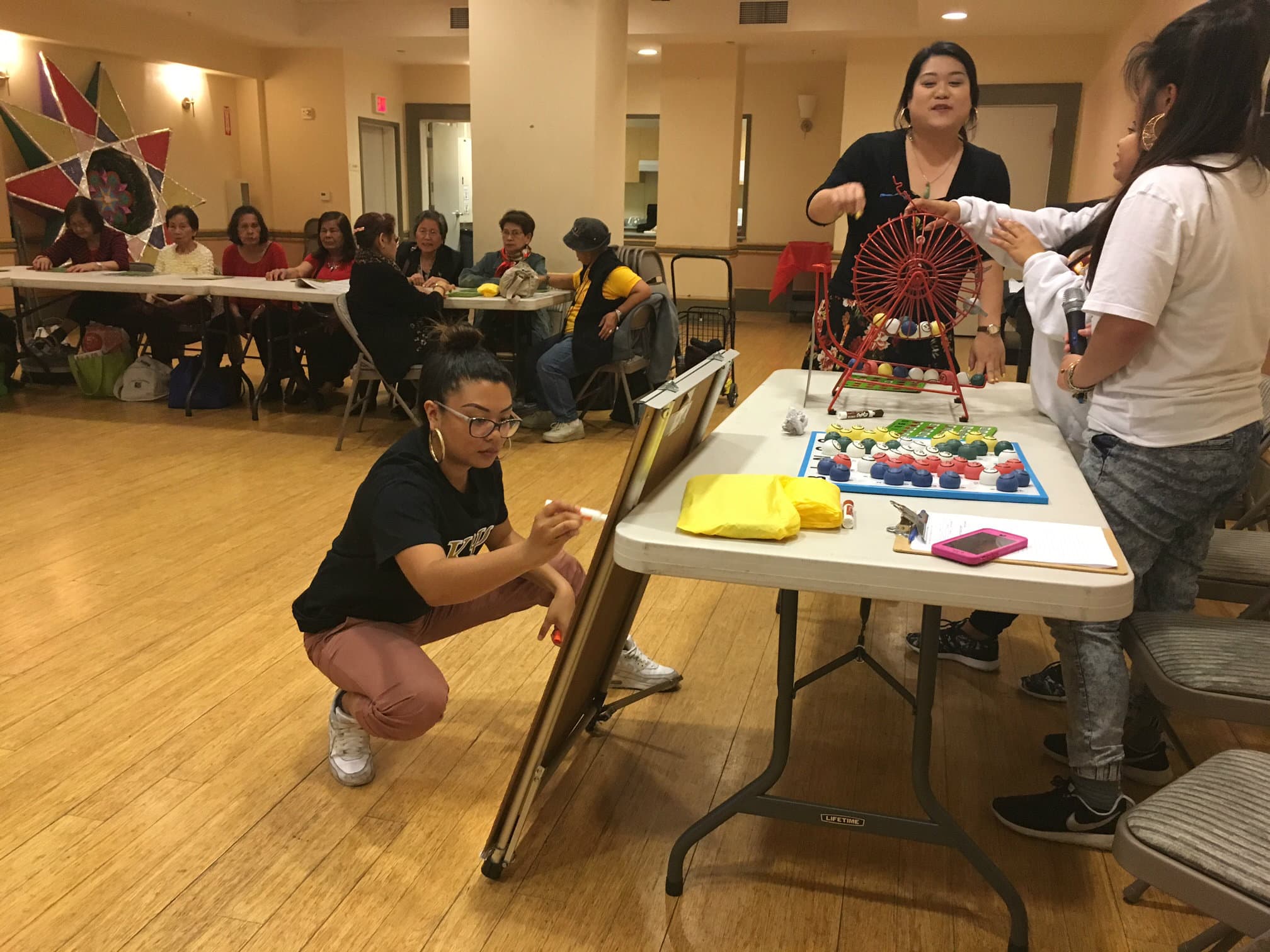 Elderly people sit at a long table, as younger people set up a game of bingo