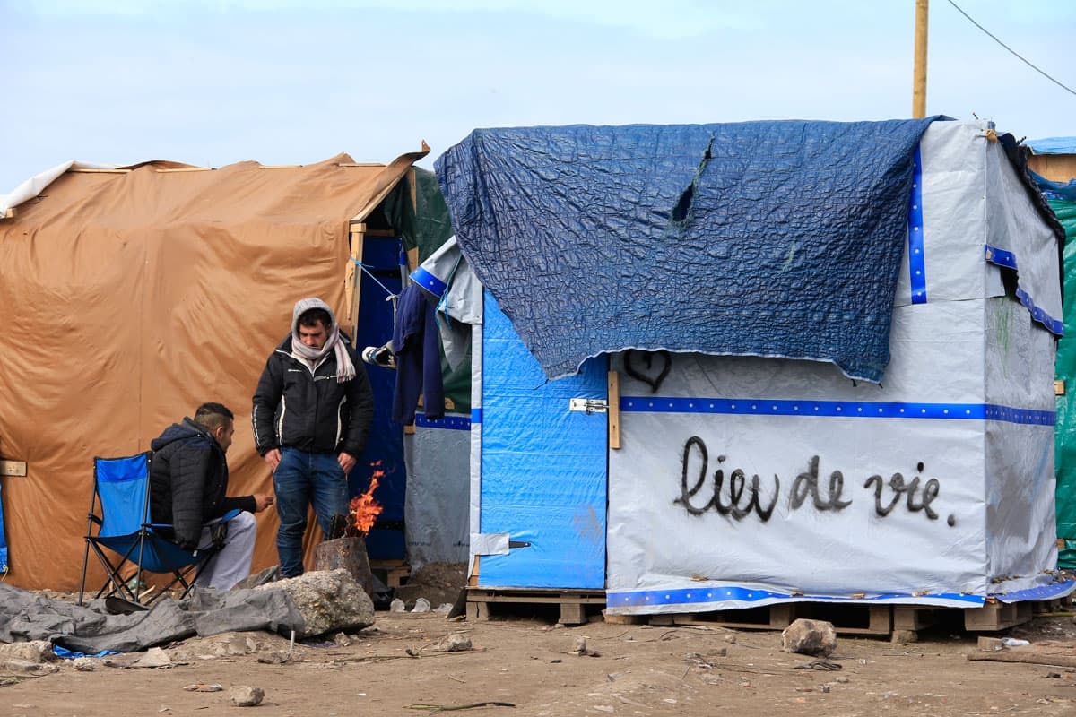 Migrants get warm near a fire outside a makeshift shelter with the message, "Place of Life."
