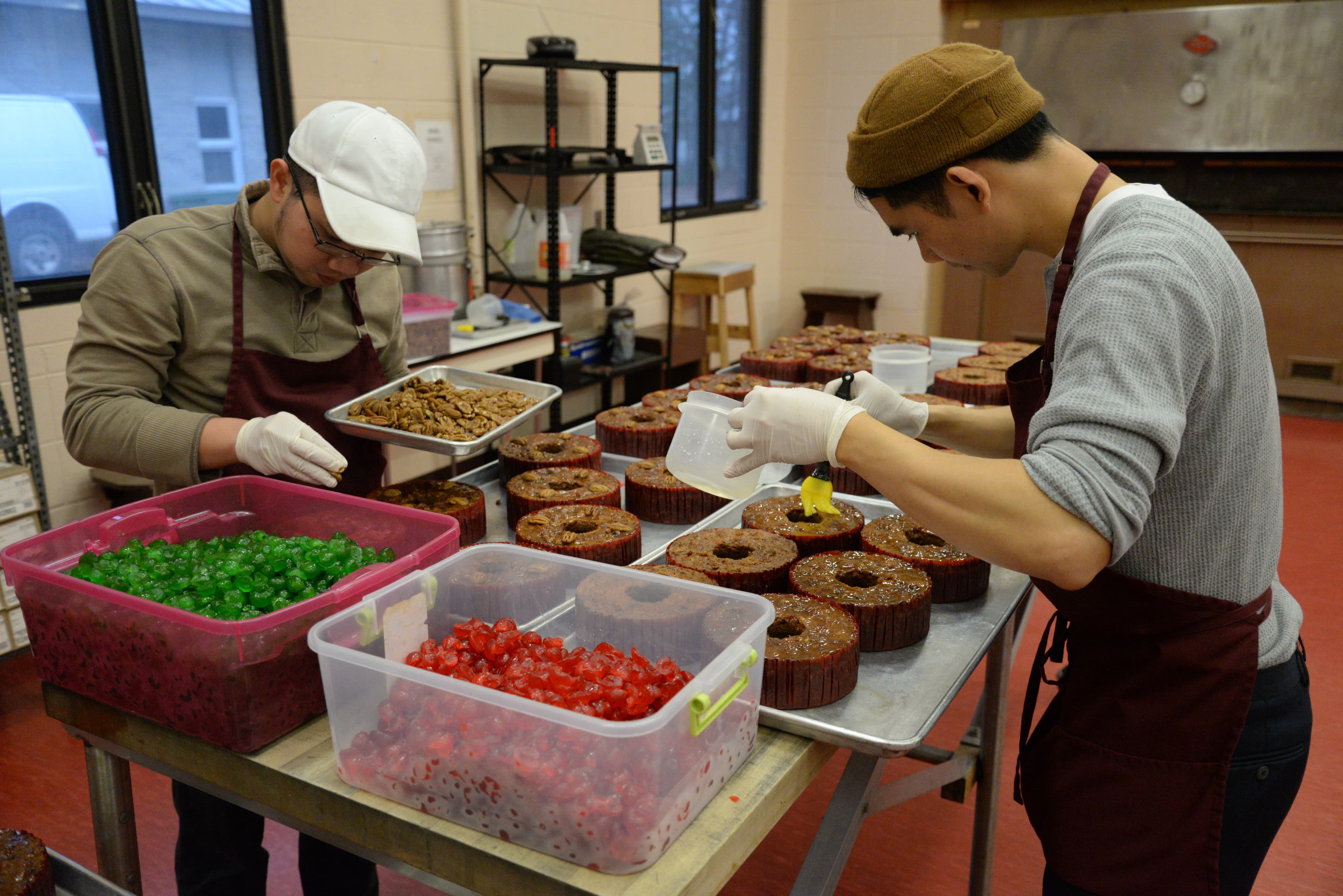 Two man stand at table with kitchen tools, over rows of brown fruitcakes