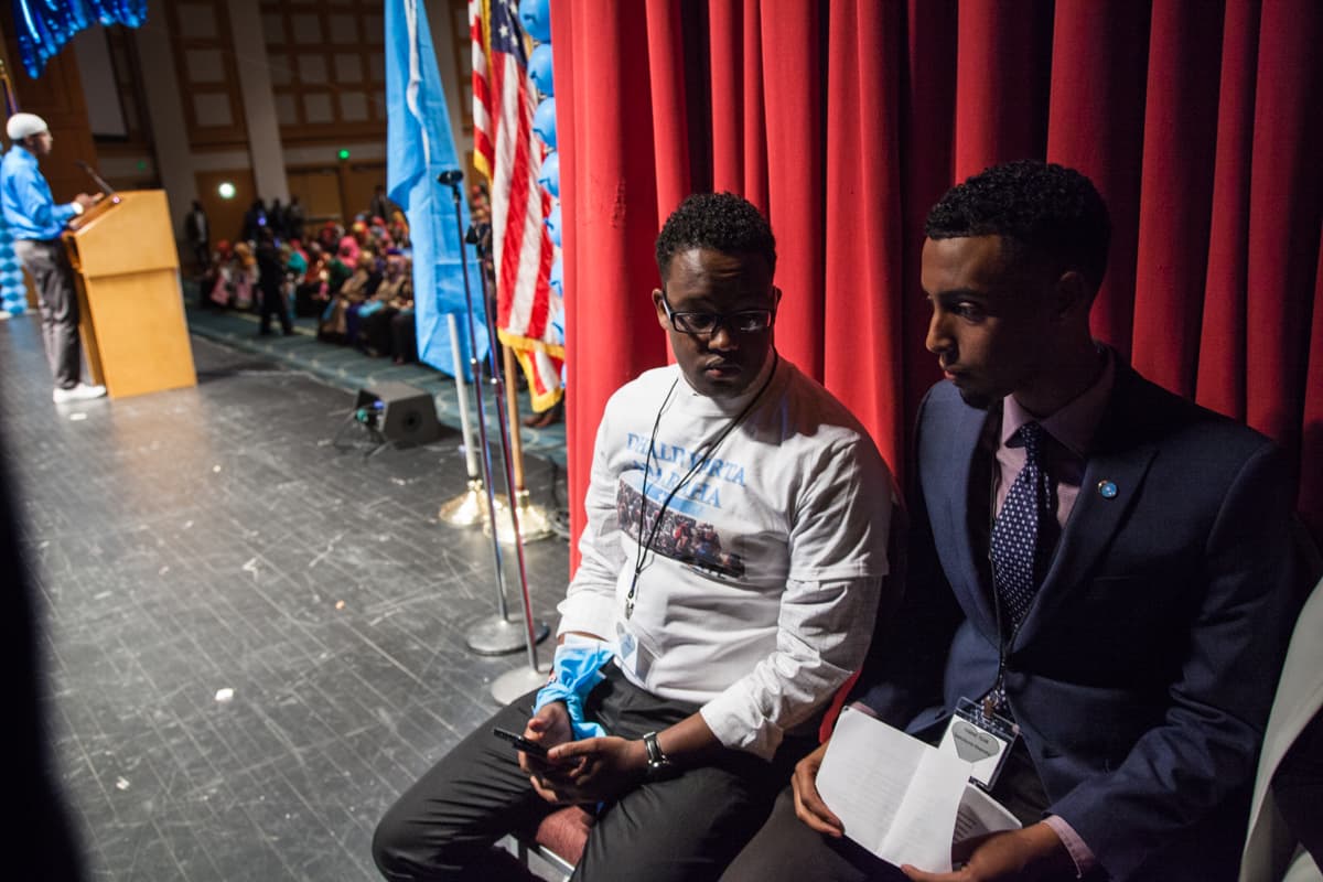 Two men sit in the wings of a stage, with audience waiting