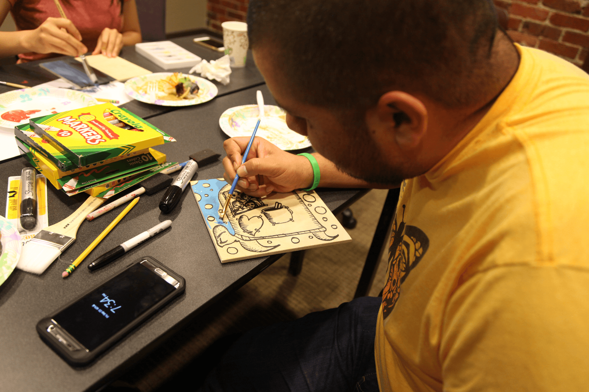 Man paints on a wooden block at a desk with others