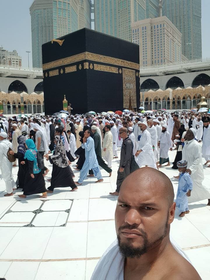 Man in robe in front of black square building, holy site, with worshippers around