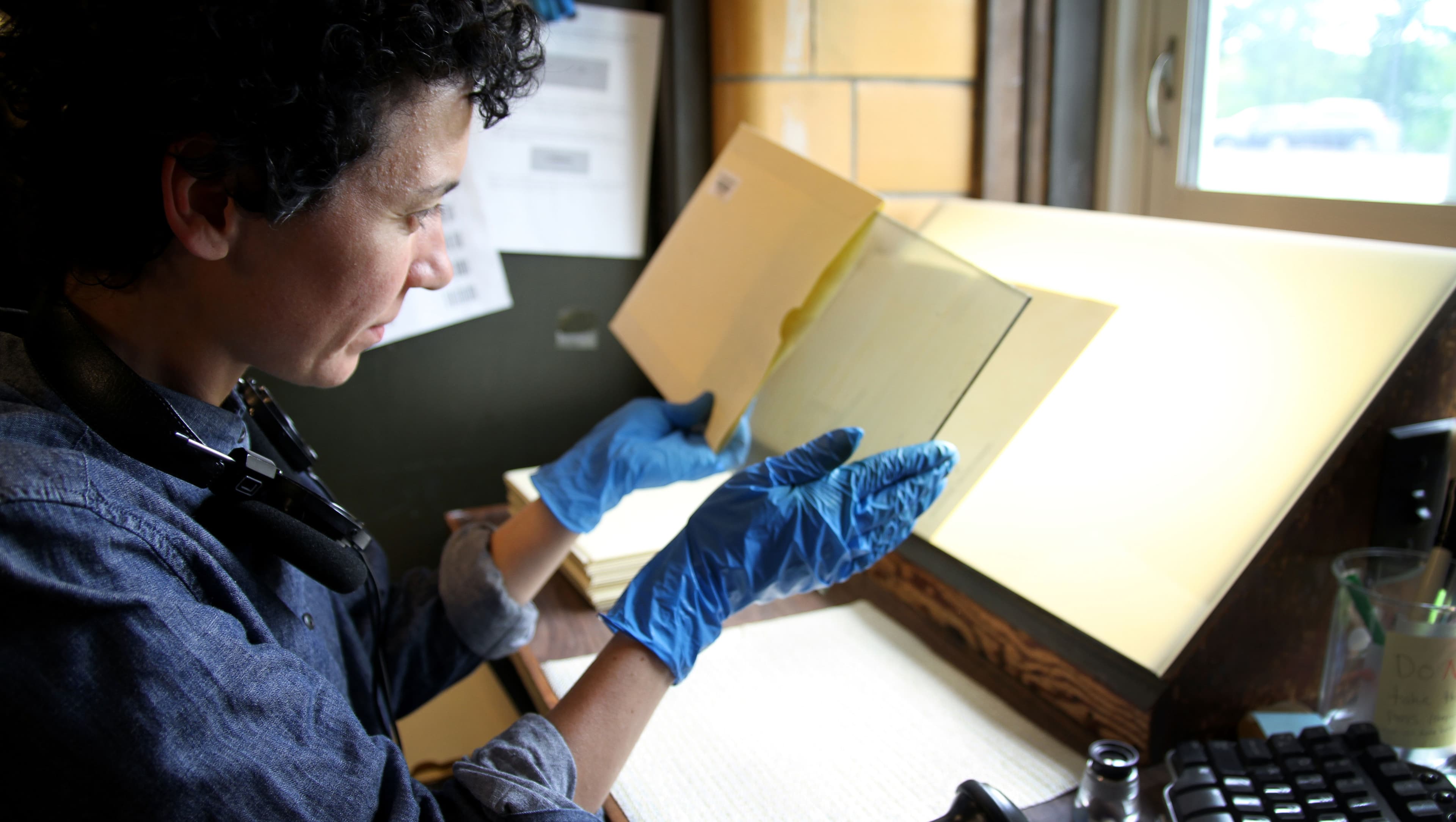 A woman holds up a glass plate, which she just removed from a paper envelope. She is staring at the plate, sitting in front of a window.