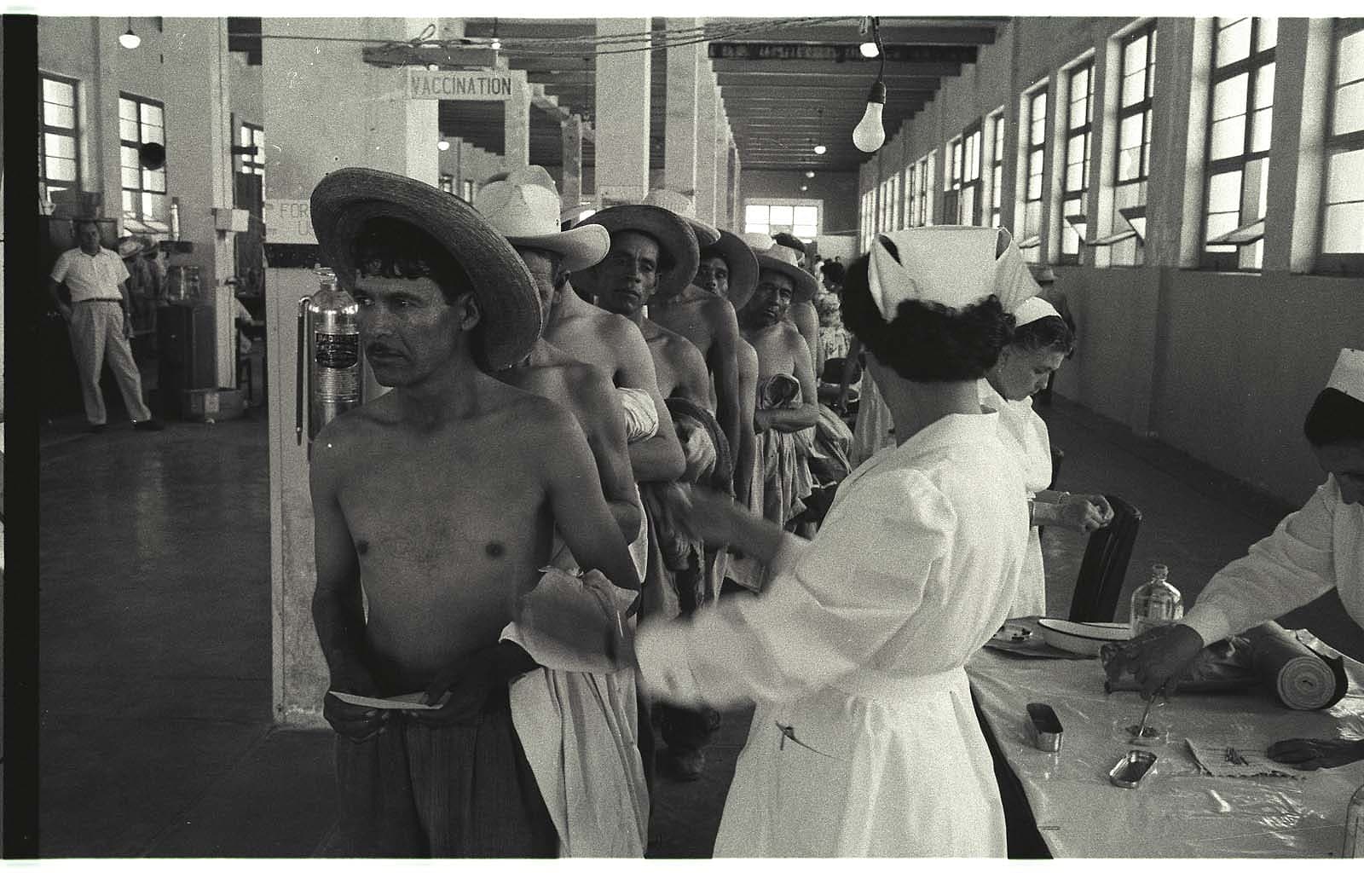 Men without shirts and wide-brimmed hats line up in front of a nurses' table.