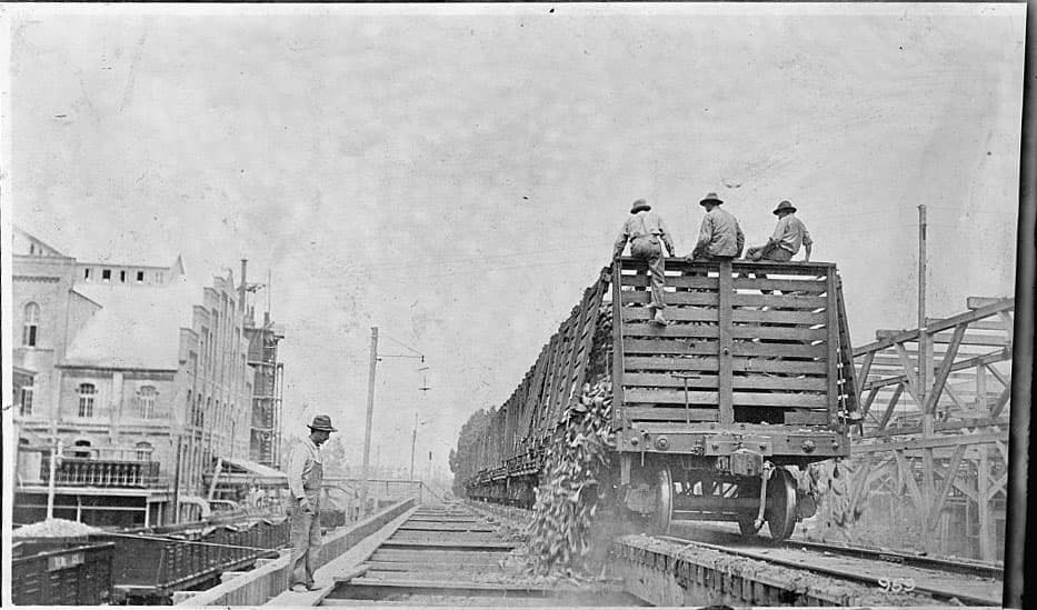 Workers sit on large crate on tracks, with vegetables spilling out, as another worker looks on. Black and white photo.