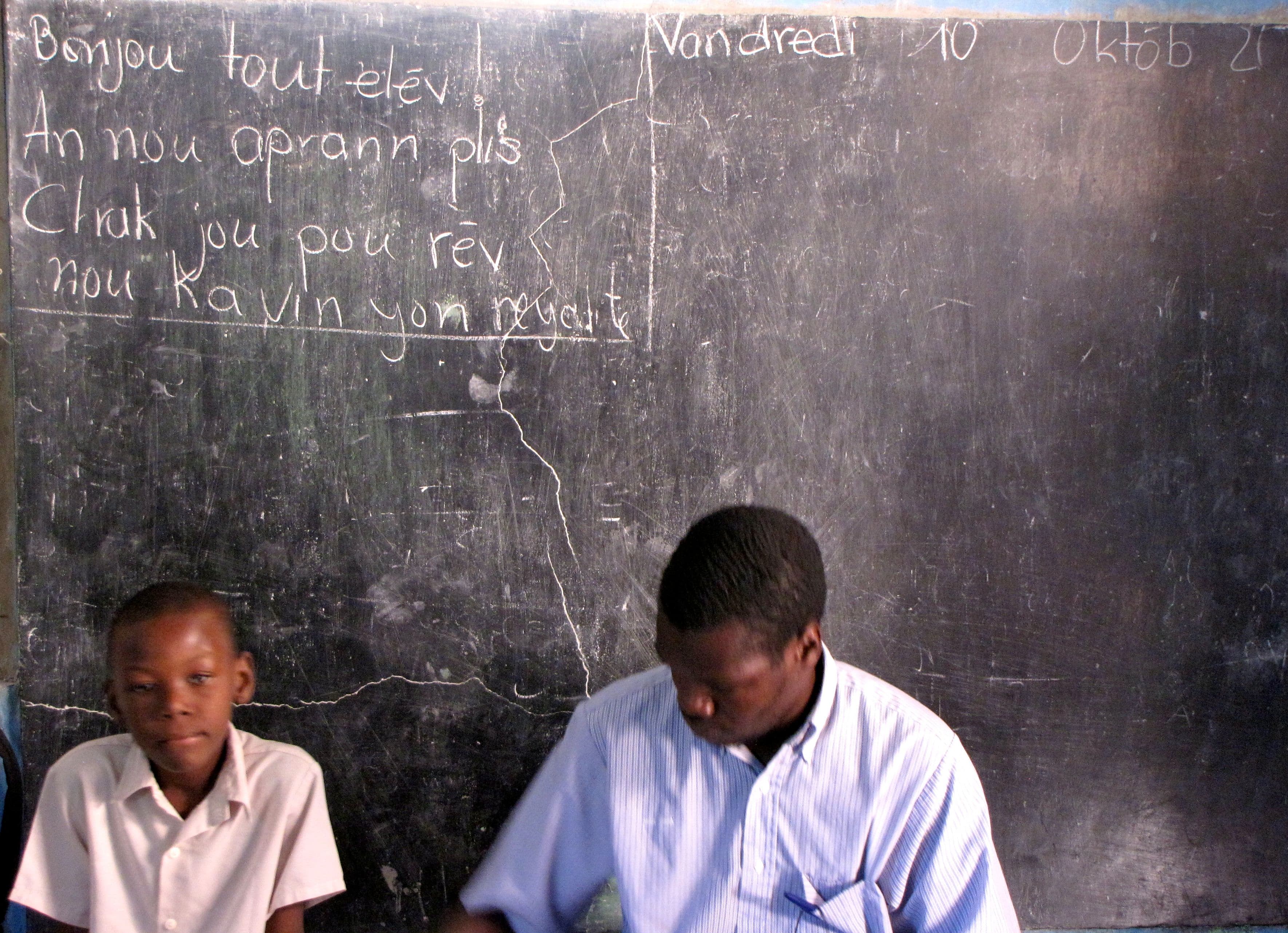 Fourth-grade teacher Kenny Antoine sits with a student. The blackboard says, 