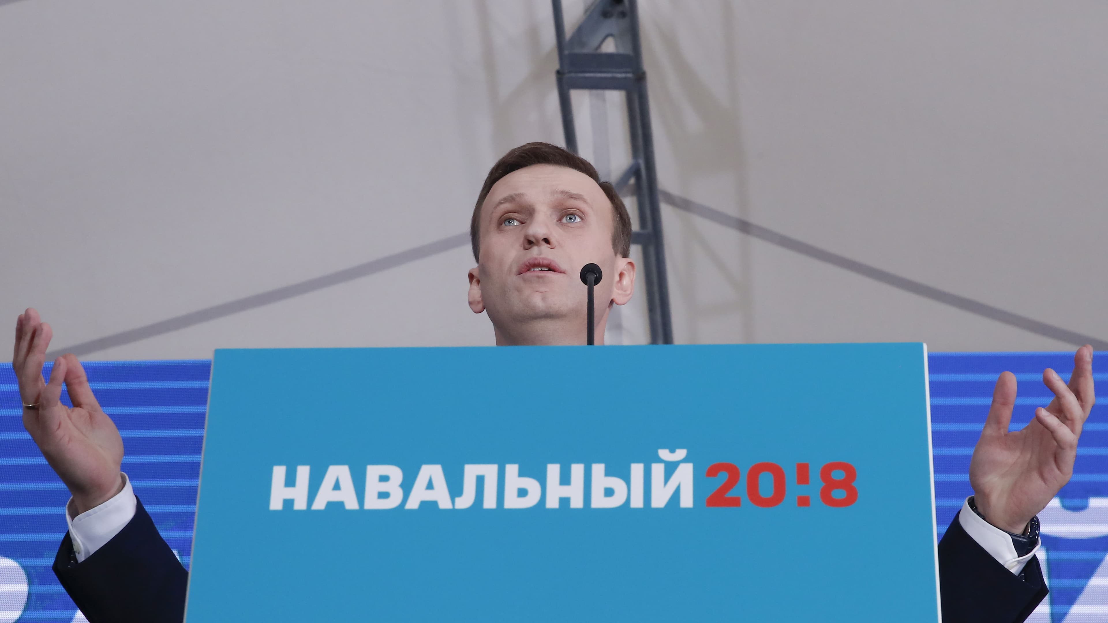 Alexei Navalny speaks behind a podium covered with a blue sign with red lettering in Russian and holds his hands up as he looks out on the crowd.