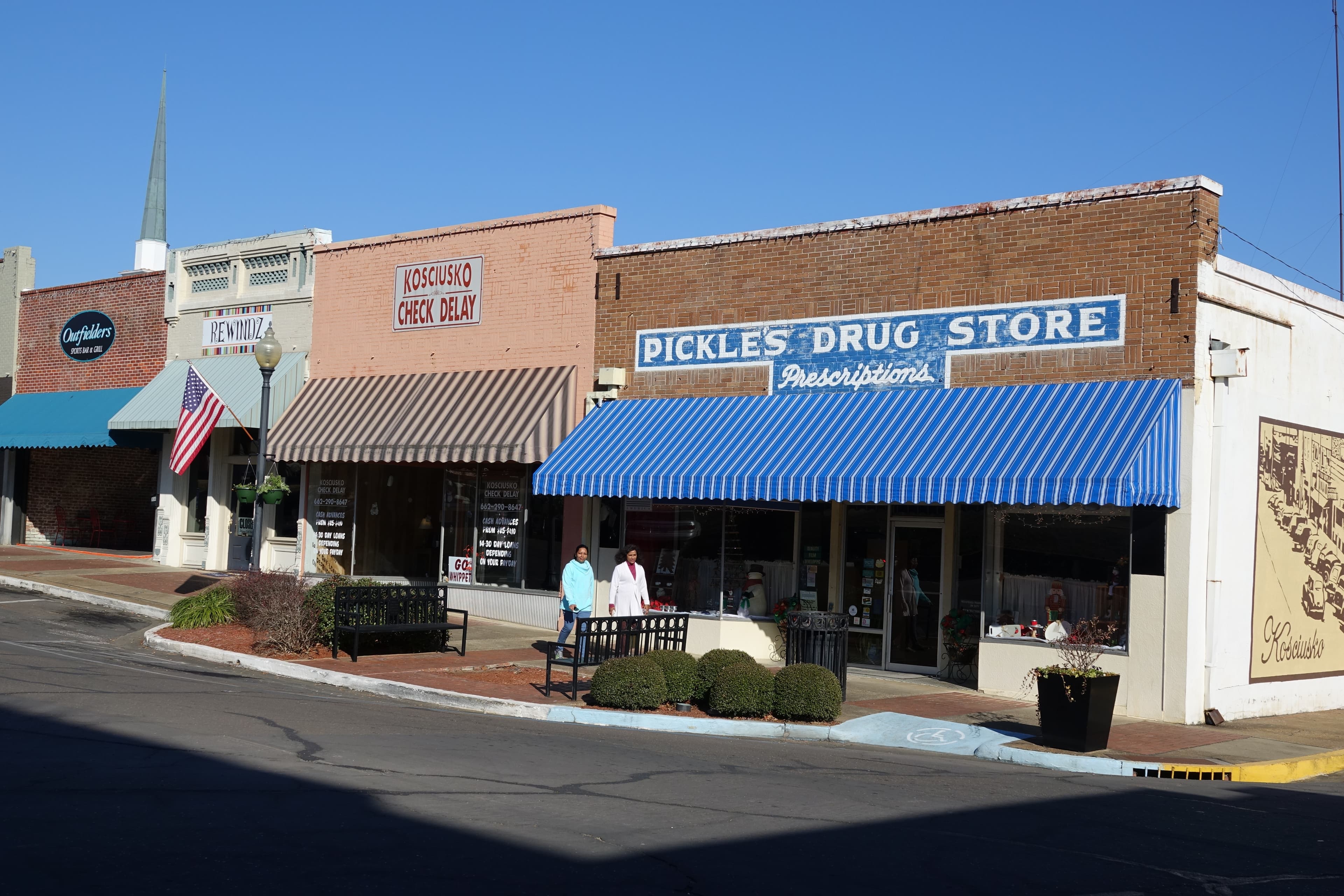 Small town road with storefronts, two women walking on sidewalk