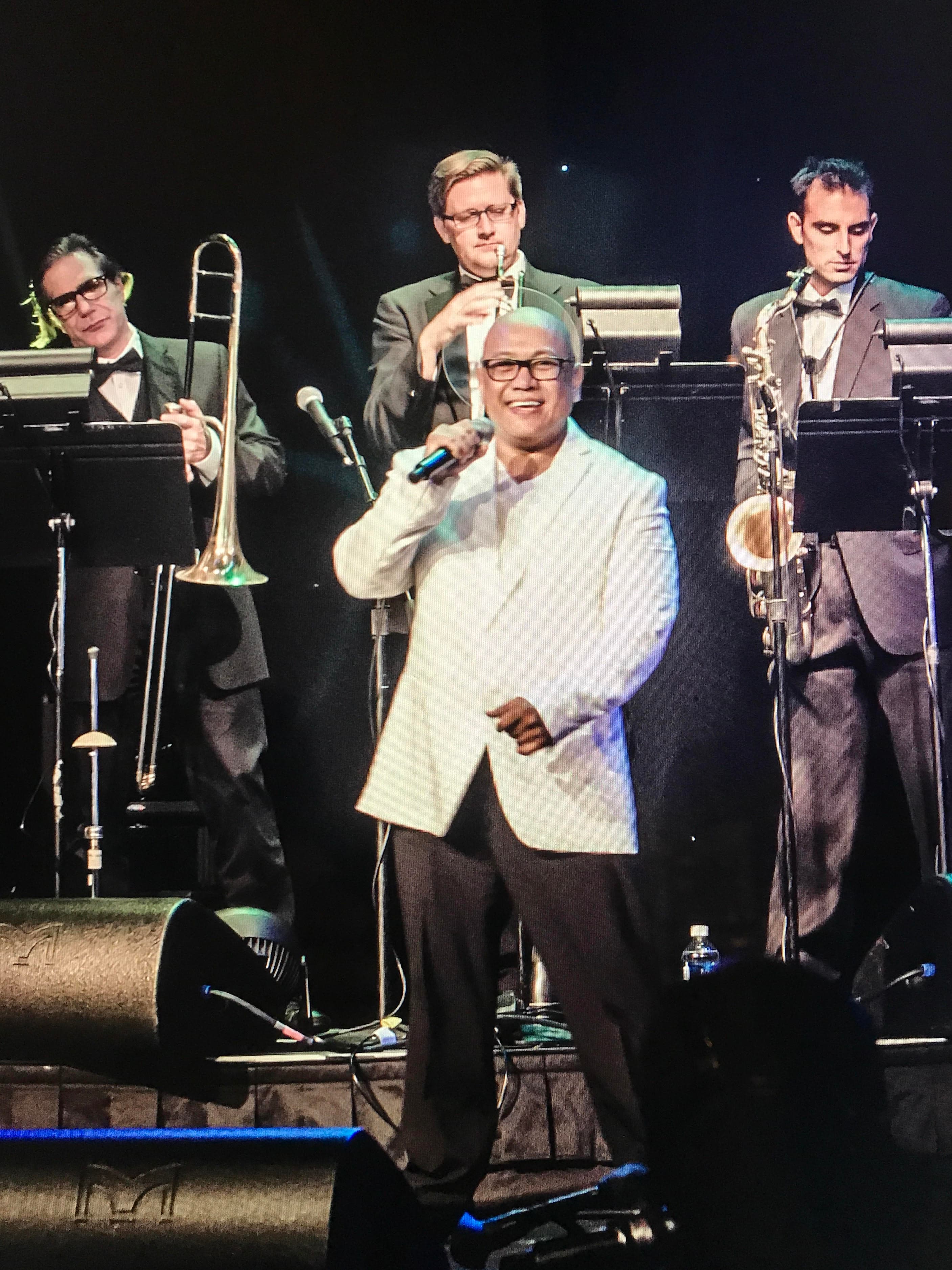 Man in white jacket holding mic on stage in front of band