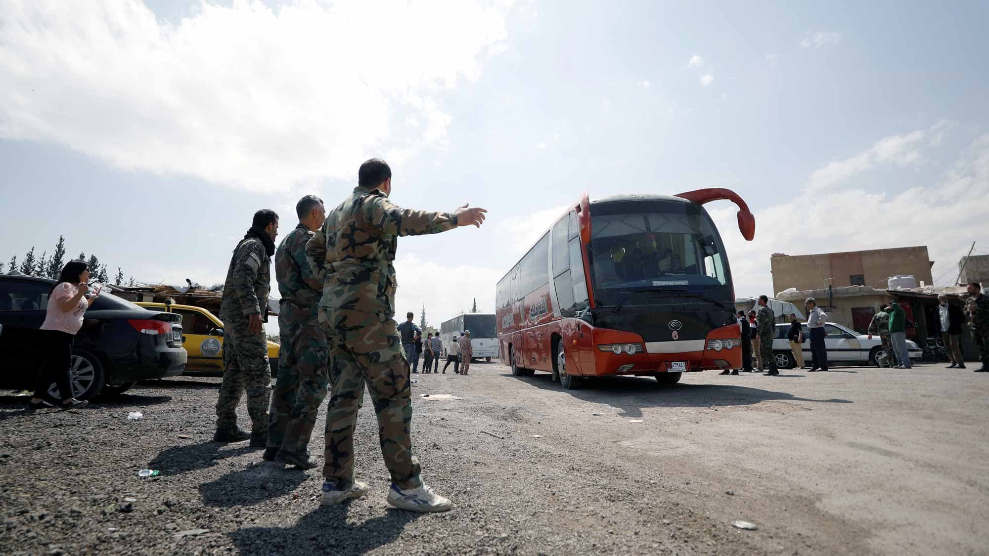 Three people in camouflage watch as a bus heads down a dirt road.