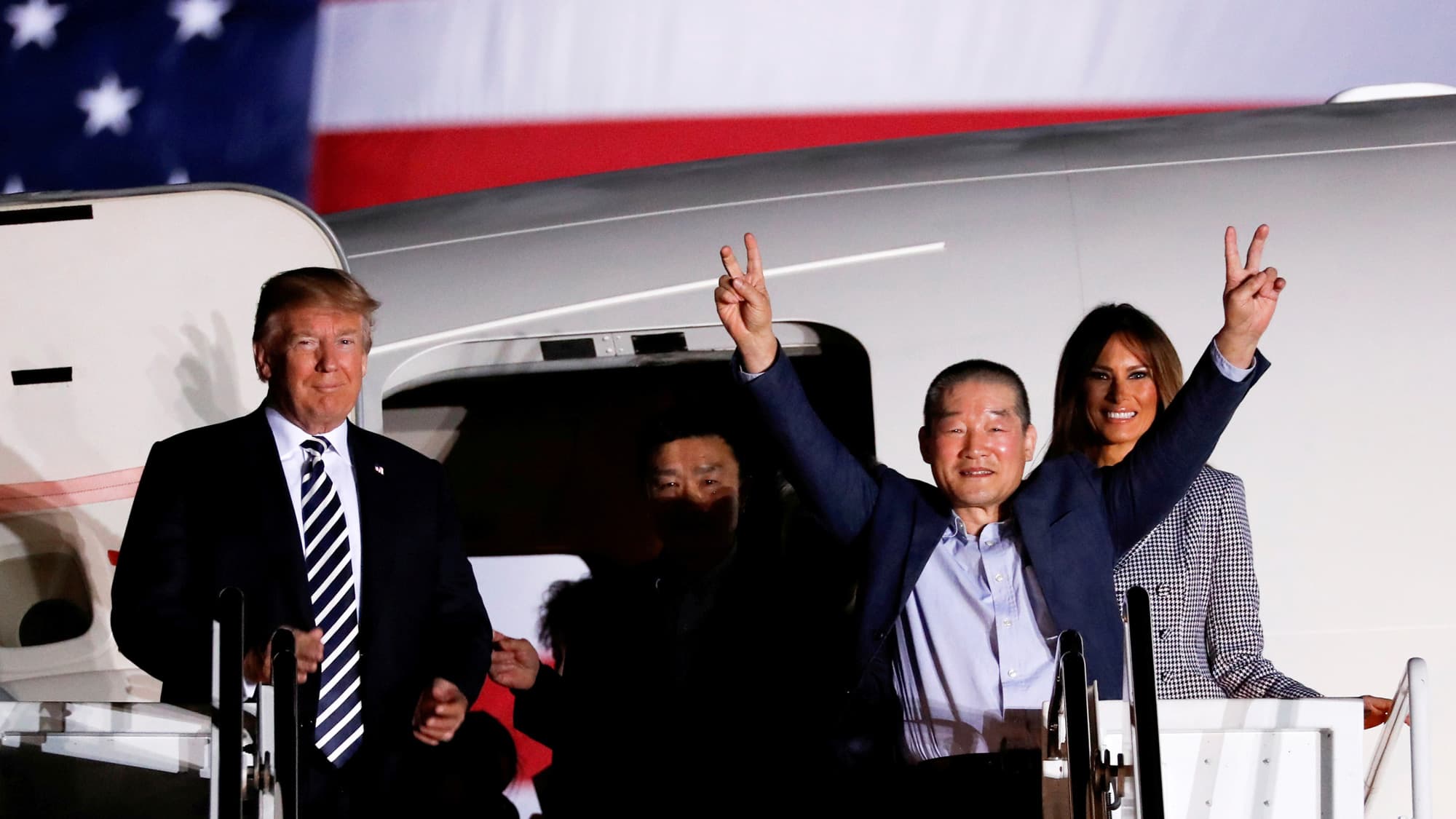 A man holds two fingers up on each hand as he exits an airplane. On the left is Donald Trump. Melania Trump is on the right.