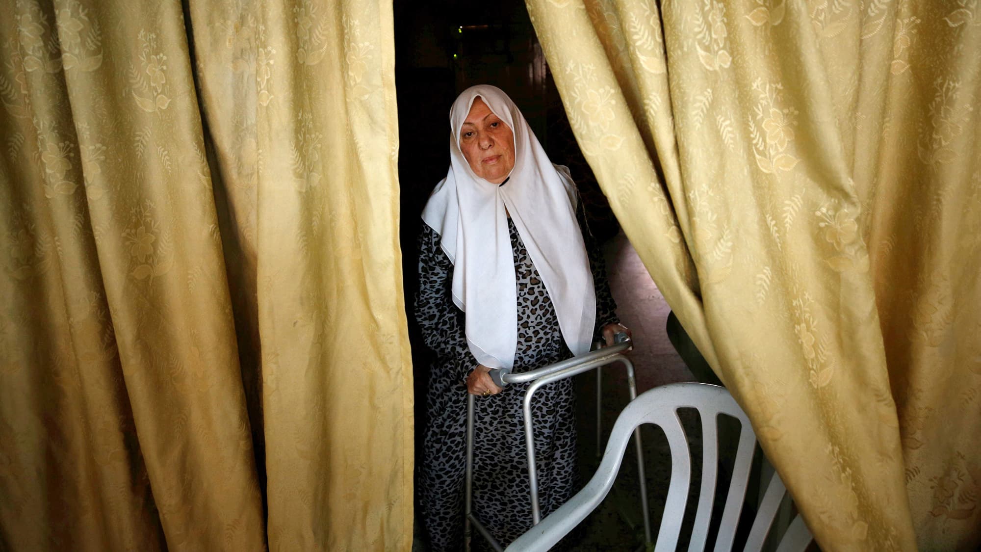 A woman wearing a white head scarf peers out from between two heavy yellow curtains. Her hands are on a walker.