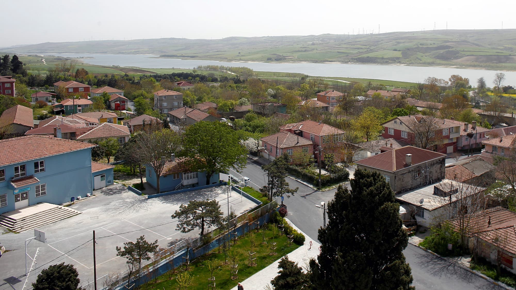 Small red homes sit peacefully next to a small, slow moving river. On the other side are gradual, green, rolling hills.