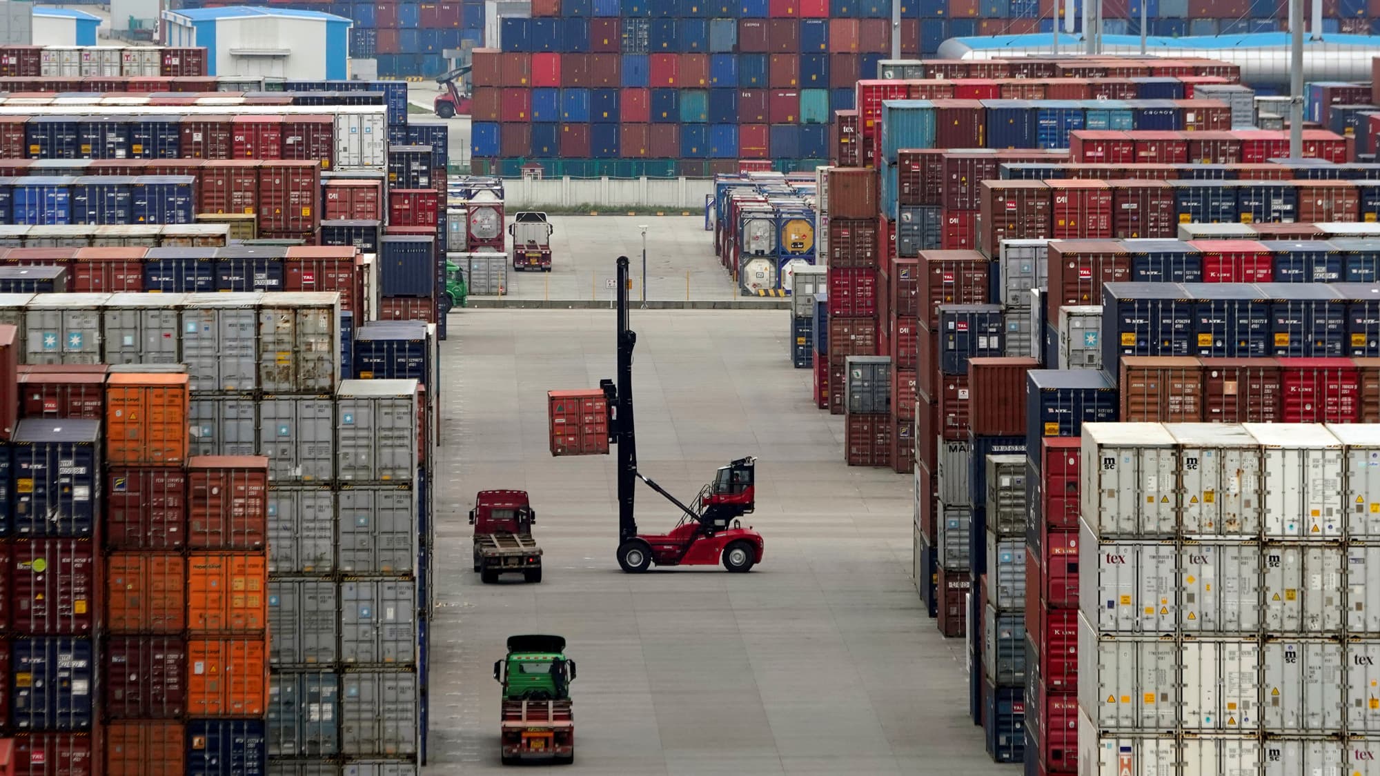 Workers use machines to move containers. The vehicles are dwarfed by tall stacks of containers — piled about six high — on all sides.