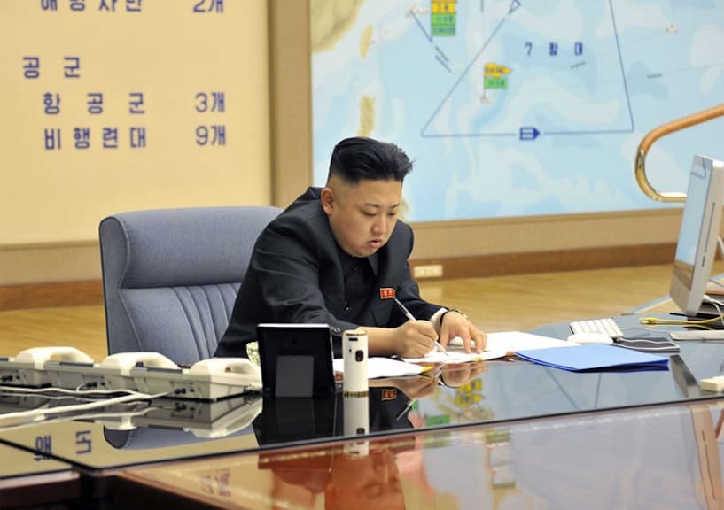 Kim Jong-un sits at a desk writing on a piece of paper. To the right of the photo you can see an iMac computer.