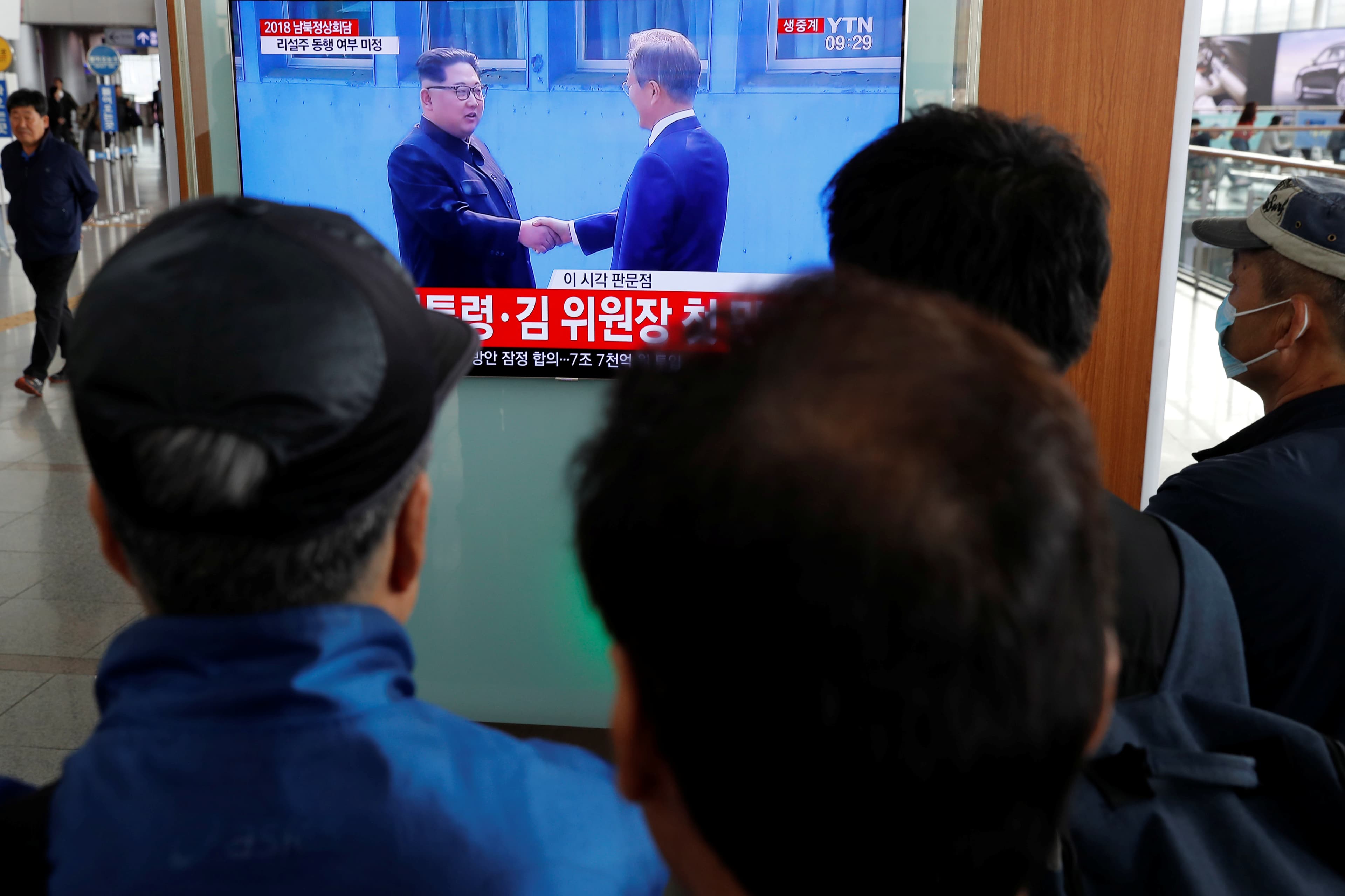 A group of people stand watching a television. Their backs are to the photographer. On the wall is a television they are looking at that shows two men shaking hands.