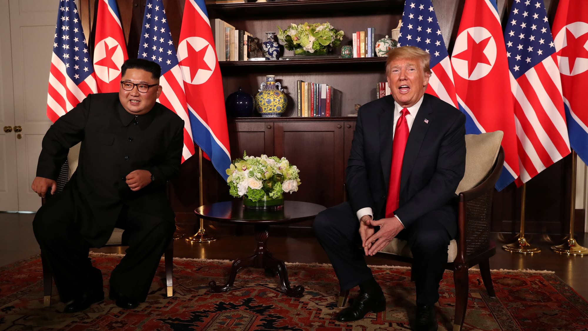 US President Donald Trump sits next to North Korea's leader Kim Jong-un with the flags of both countries behind them.