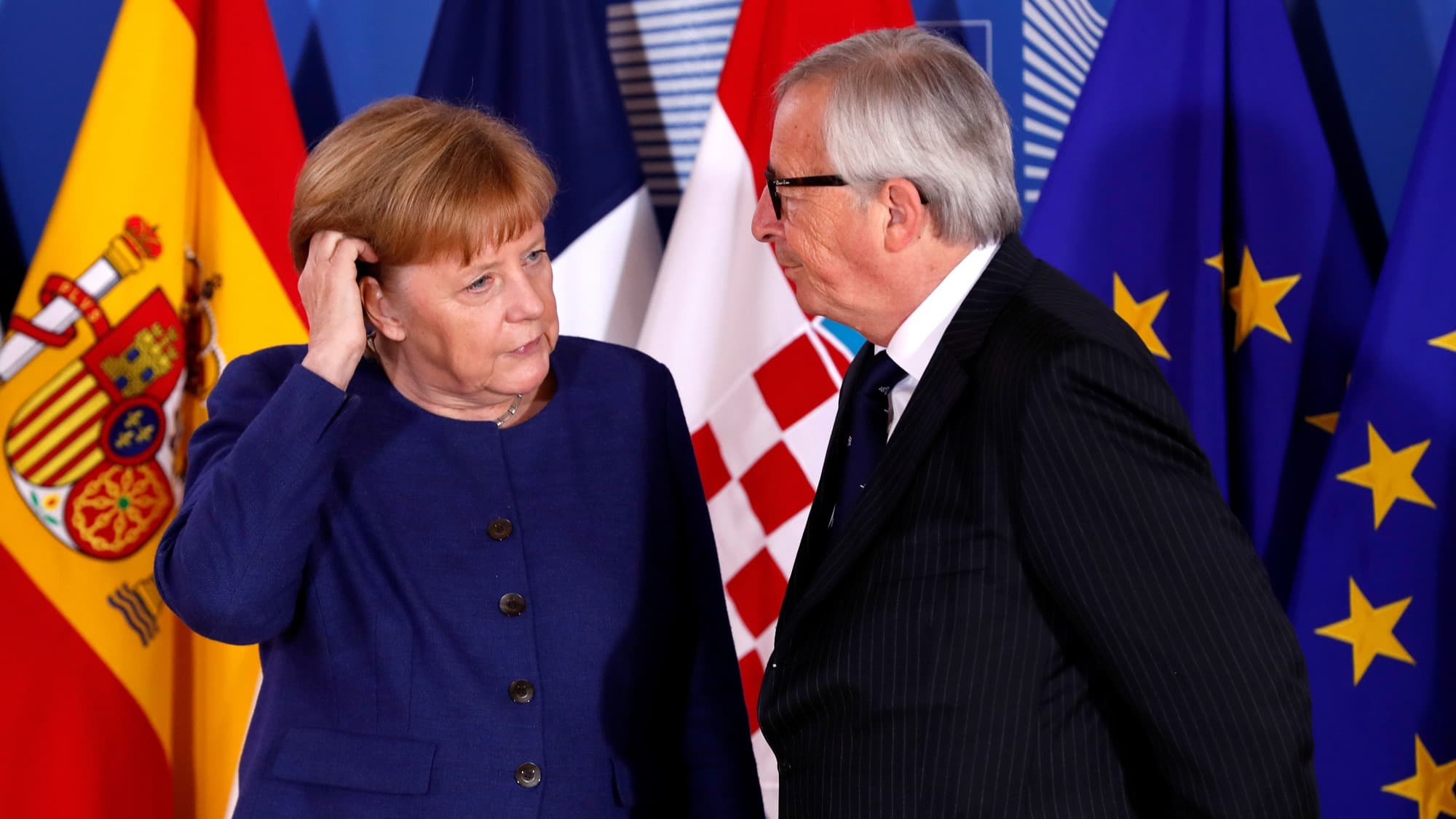 German Chancellor Angela Merkel is seen in a blue dress next to European Commission President Jean-Claude Juncker in a blue suit with several flags behind them.
