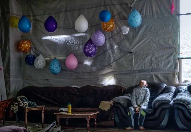 A young boy sits on a couch, looking toward the ceiling, where a string of balloons hangs