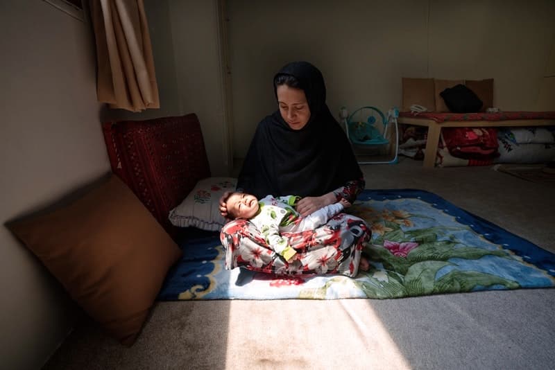A woman sits on the ground and holds a baby in her lap. Sun shines on her in a room that is otherwise dim.