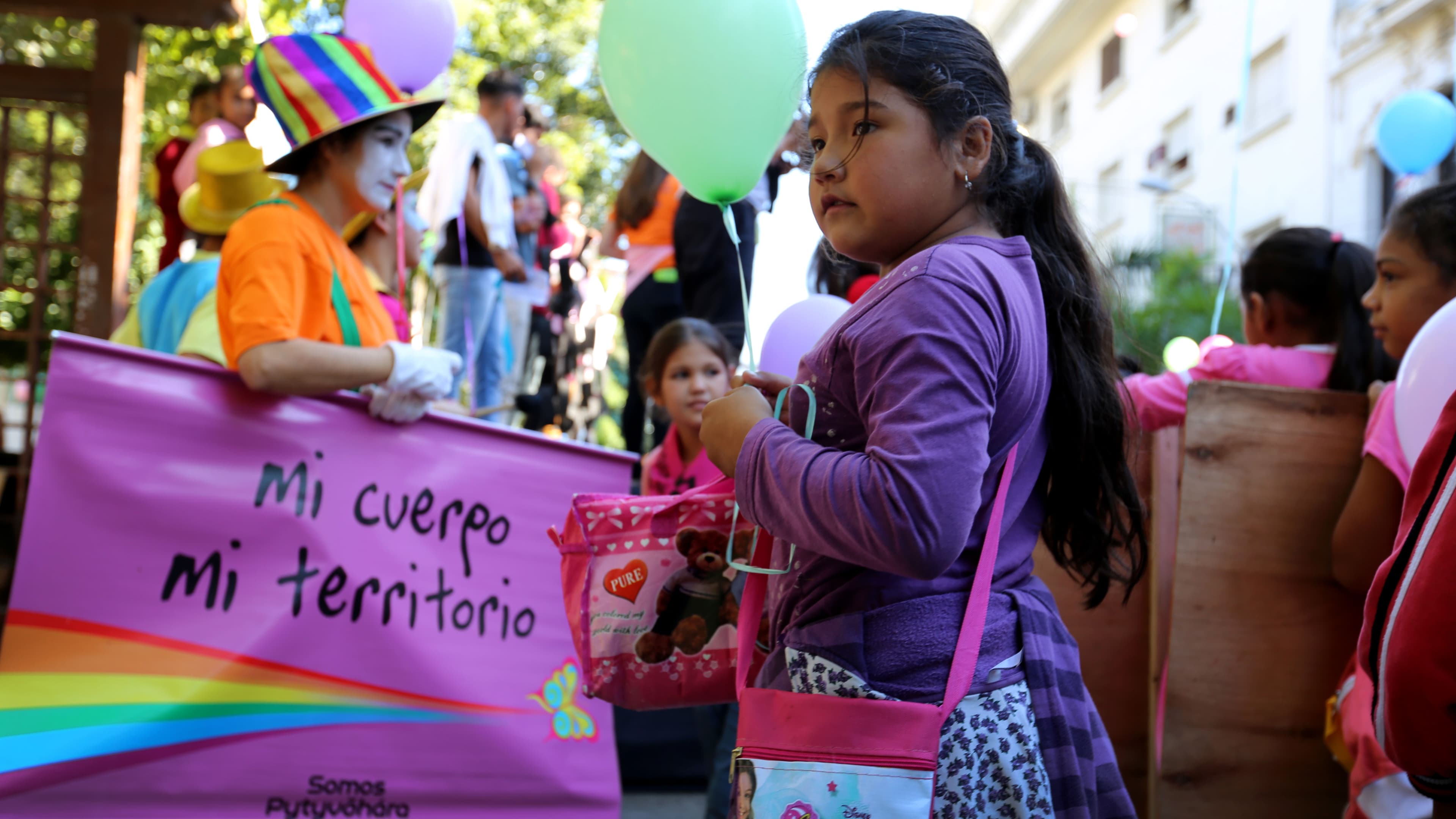 A young girl holds a balloon. Behind her are protestors carrying signs and children with more balloons. One person is dressed as a clown with a sign that says, "my body, my territory."