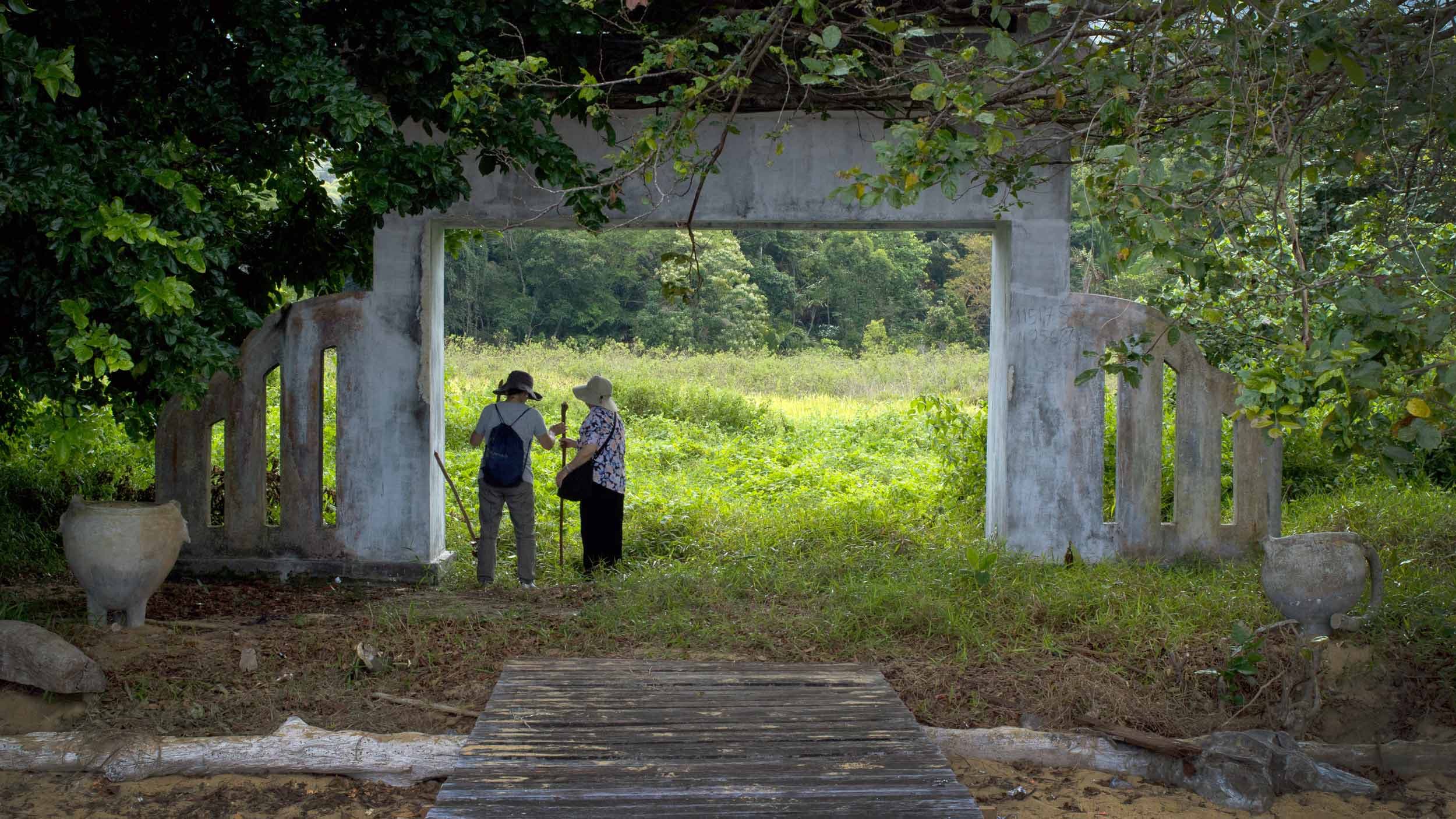 Two people in large hats stand under a stone arch that is covered by leafy foliage.