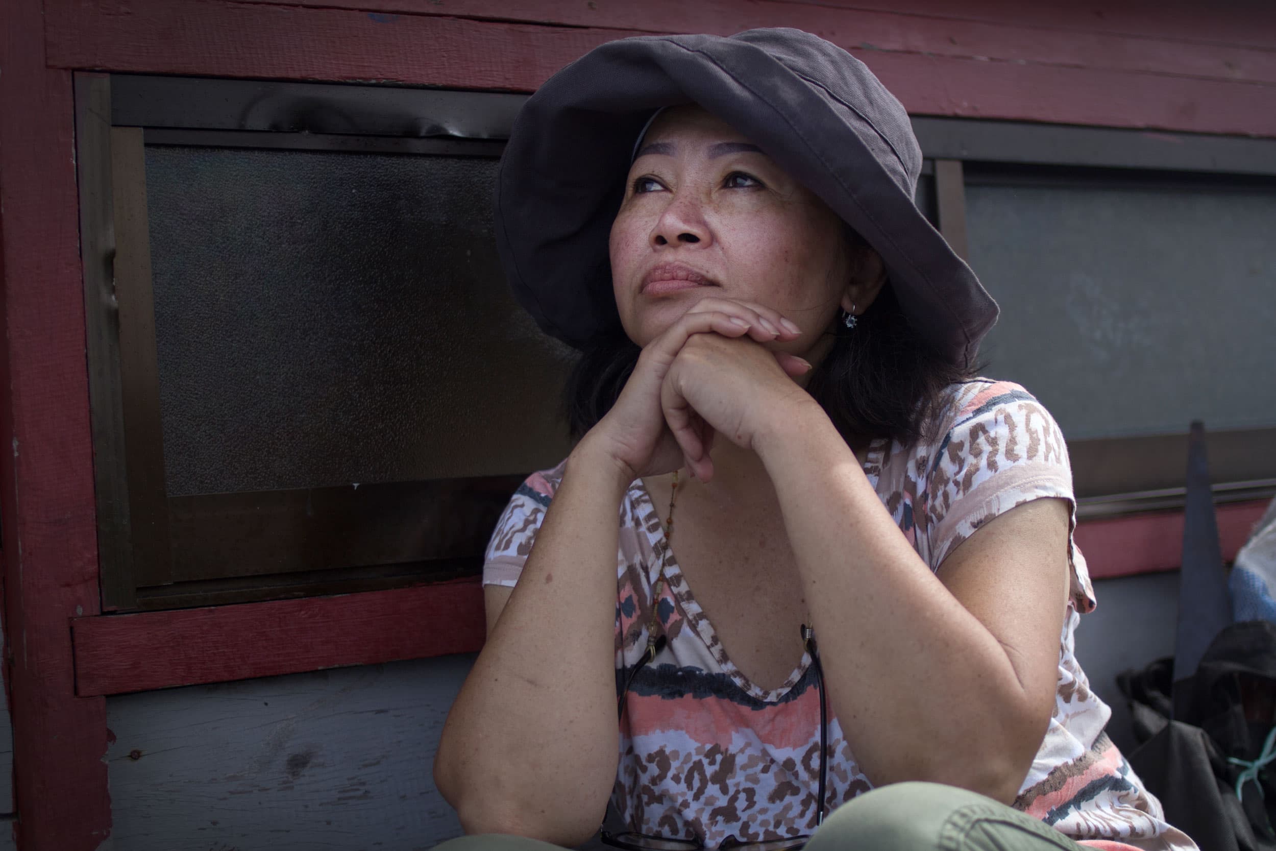 A woman wearing a brown hat sits against a wooden wall with her hands folded under her chin