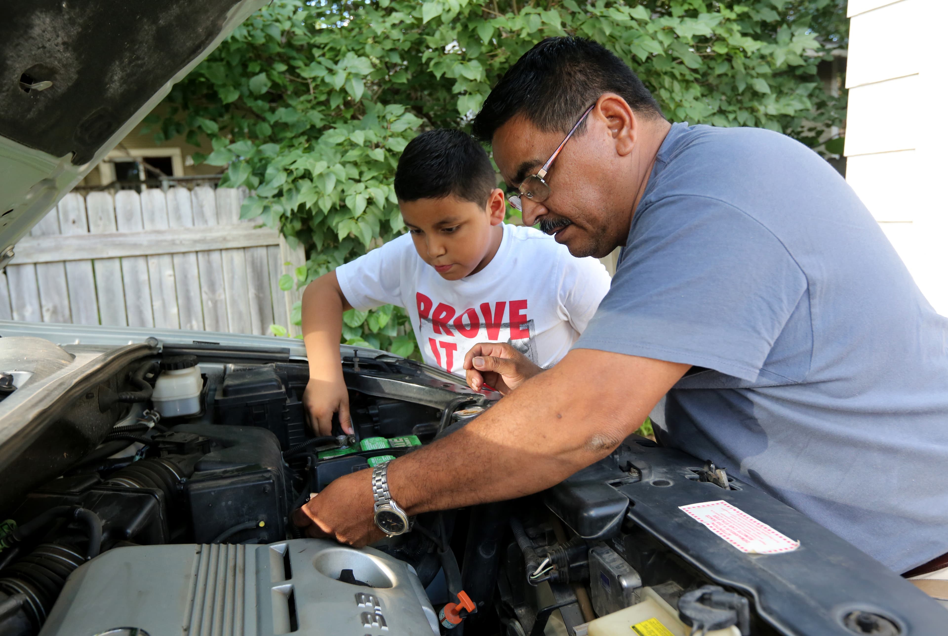 Man leans over open hood of car, young man standing behind him