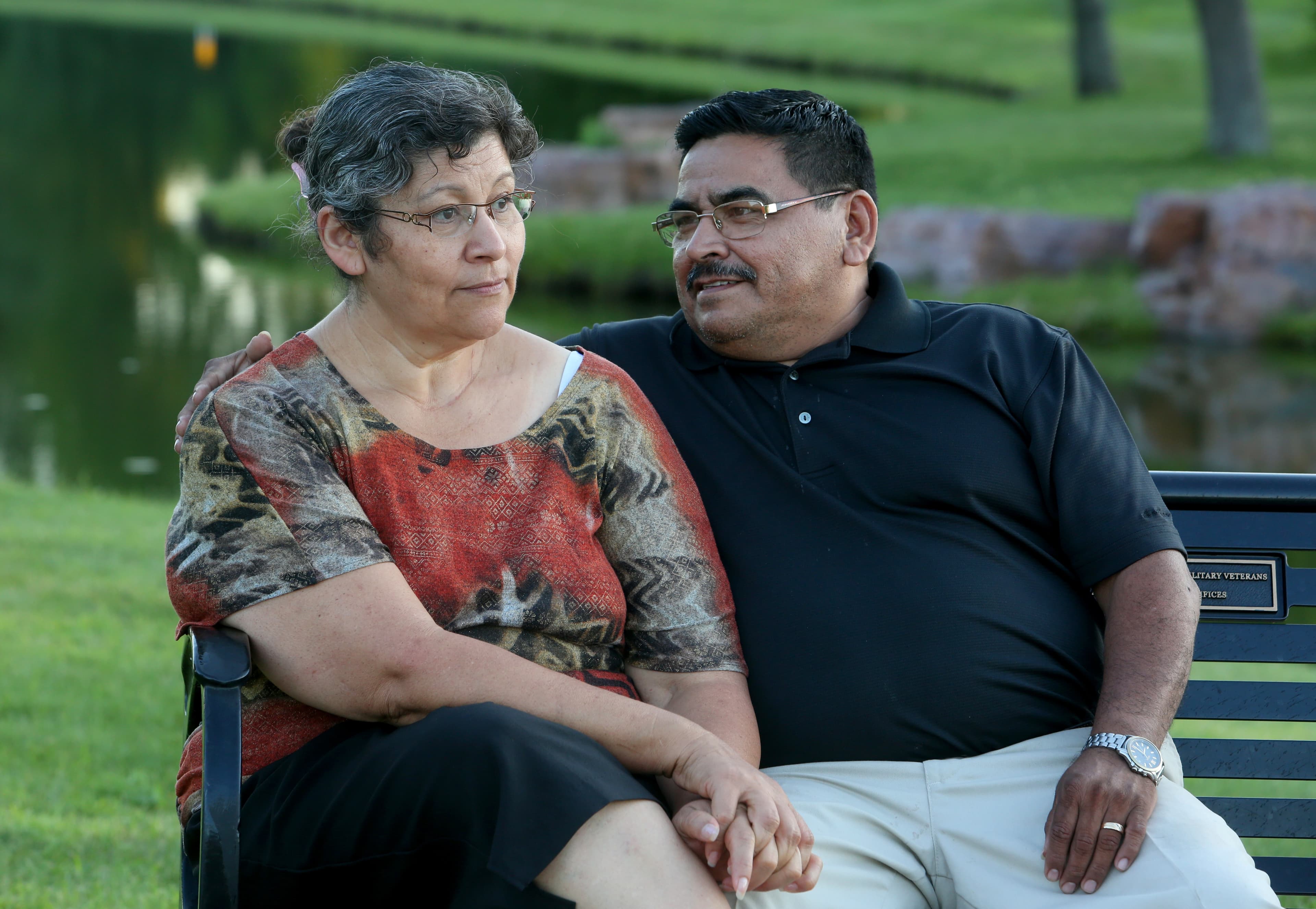 Woman and man sit on bench in front of lawn, trees. Man looks at woman with a slight smile while woman looks off into distance