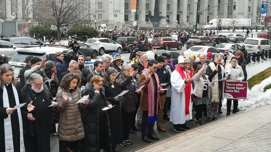 Group, including priest, lined up on street with immigration signs