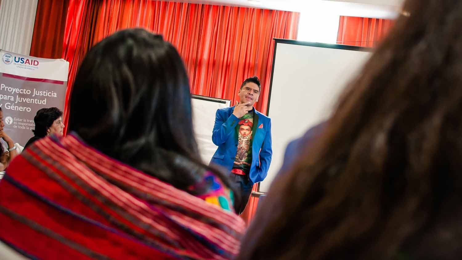 A man in a bright blue coat is speaking in front of two white screens as the audience watches him.