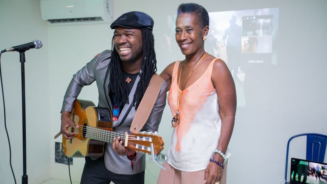 Man playing guitar in front of projected images laughing, with woman next to him, arm around his shoulder. Laptop on chair in background.