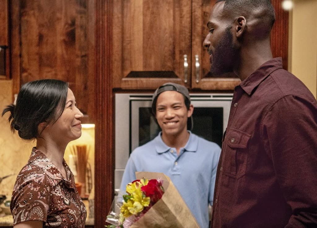 In front of wodden kitchen cupboards, older Vietnamese woman speaks to young black man
