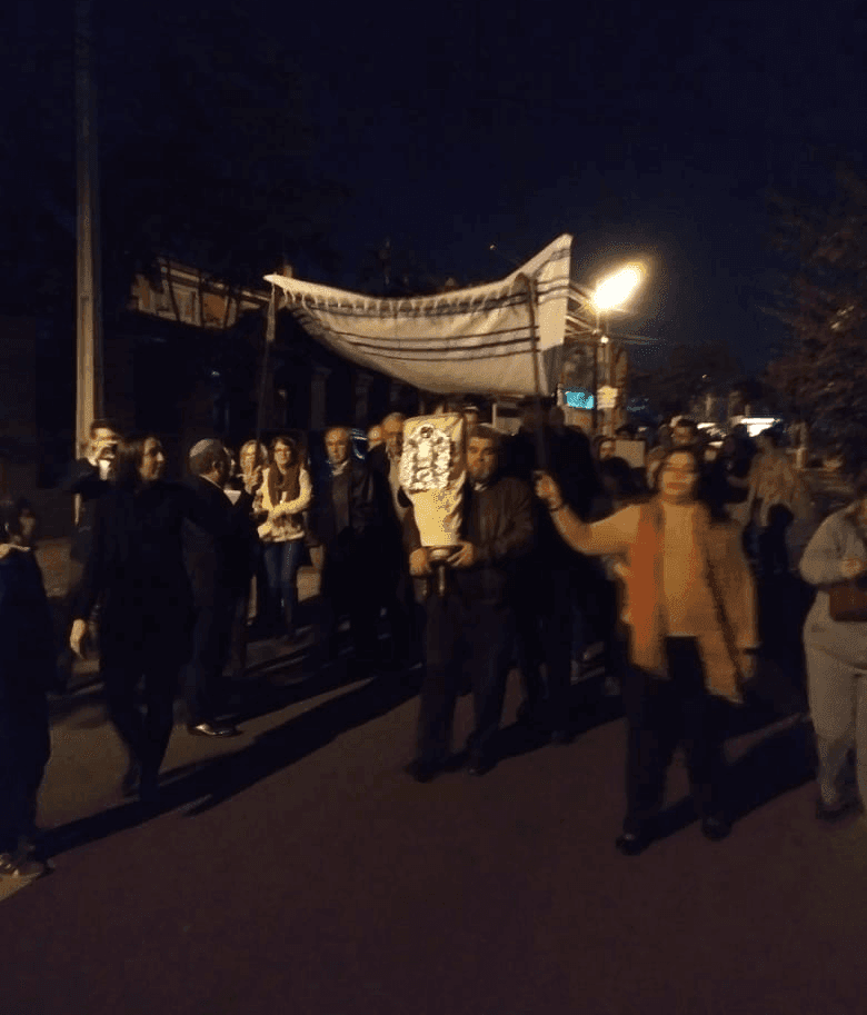 Small group walks up street holding relic, with white sheet on sticks above