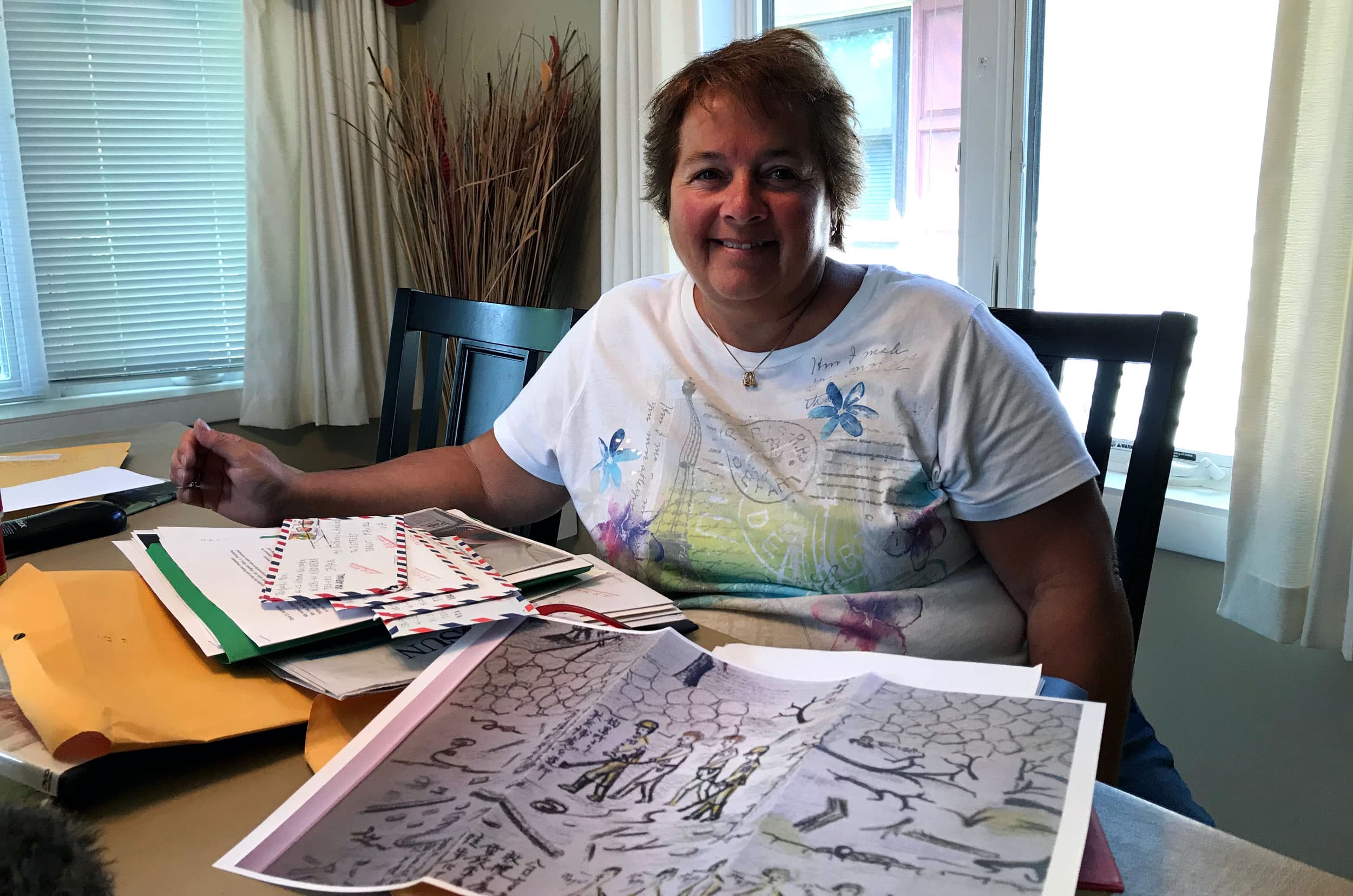 A woman sits at a table with a drawing and some letters piled in front of her.