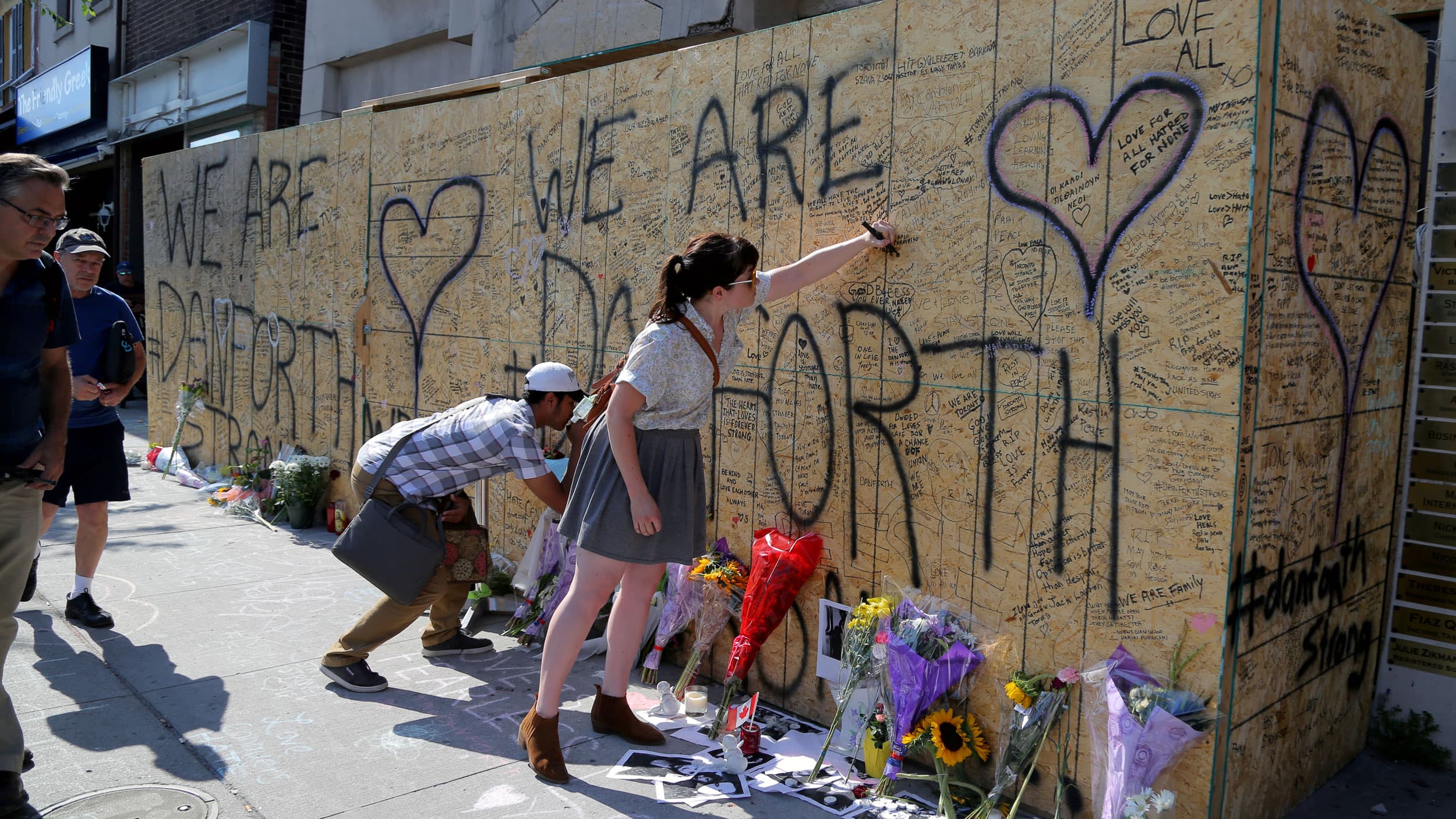 People leave messages, flowers and other trinkets on a plywood wall that says "We are Danforth"