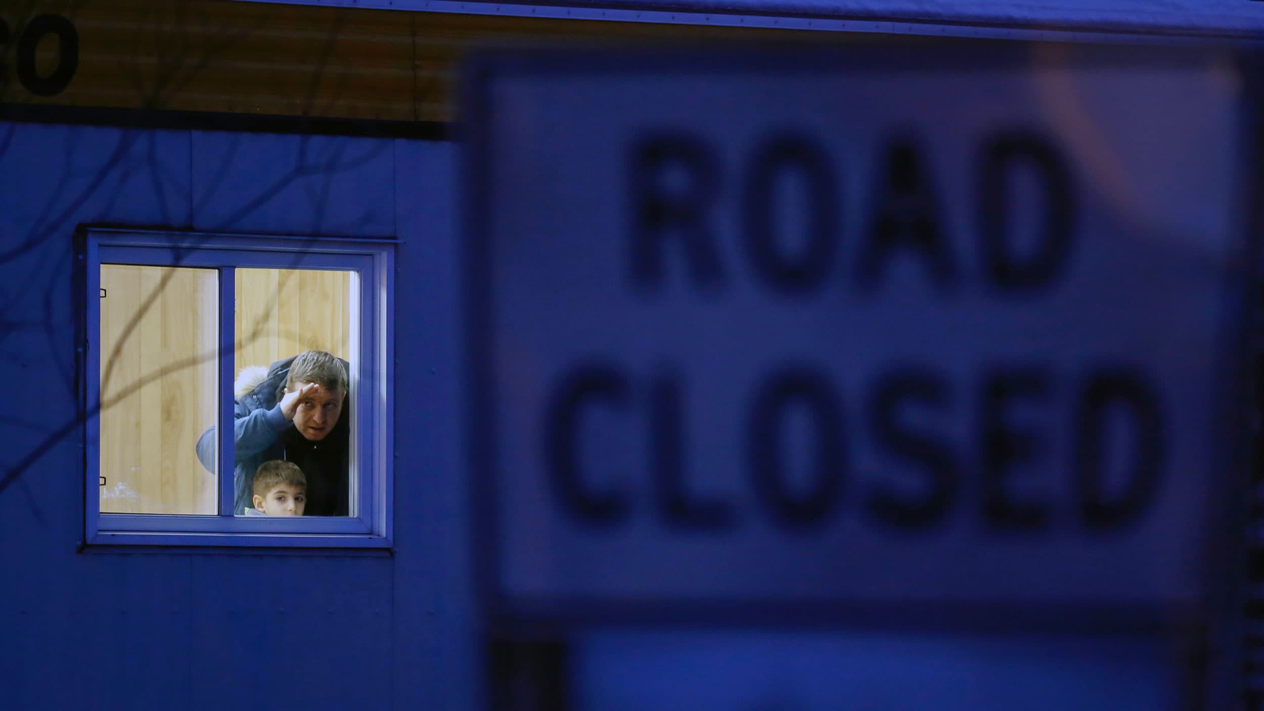 A man and a child peer out a window of a building. Next to them is a road sign that says "road closed"