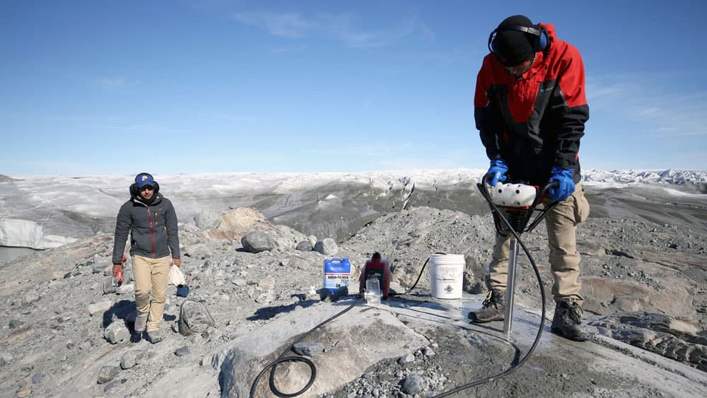 Two men stand on rocky land as mountains and snow stretch out behind them. One holds a drill of some kind and is drilling into the rock.