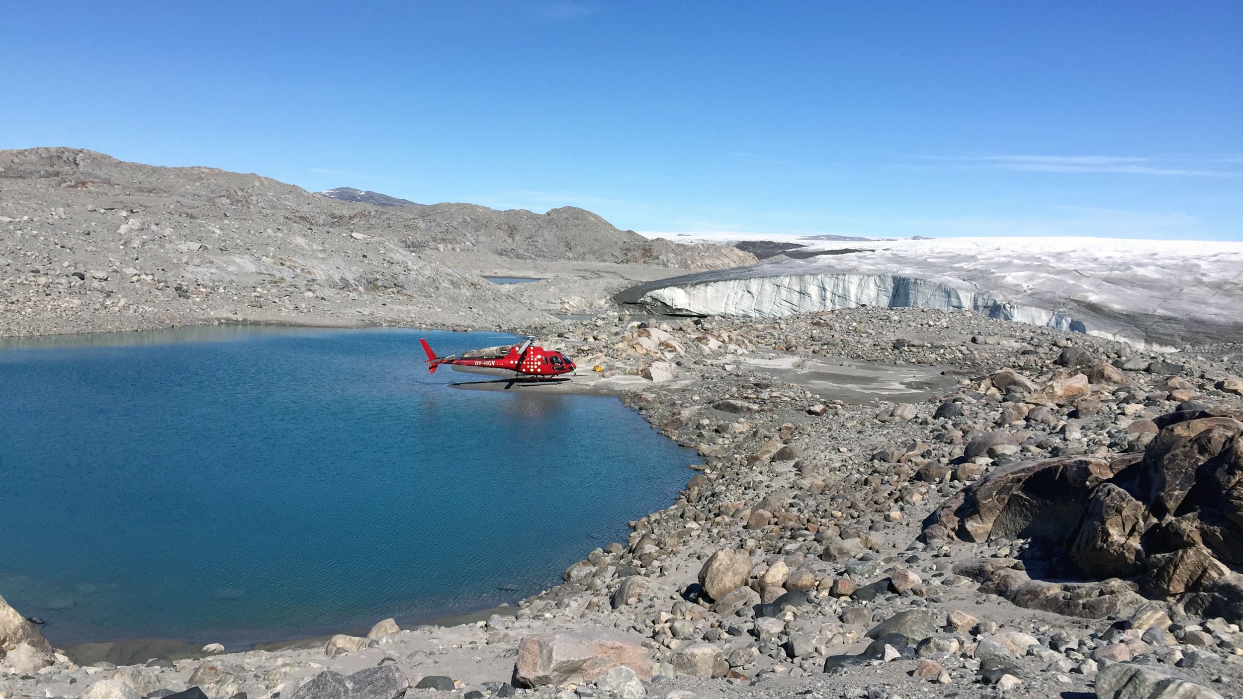 A red helicopter is landed on the edge of a blue lake on a rocky shoreline. Hills in the distance are covered in white ice.