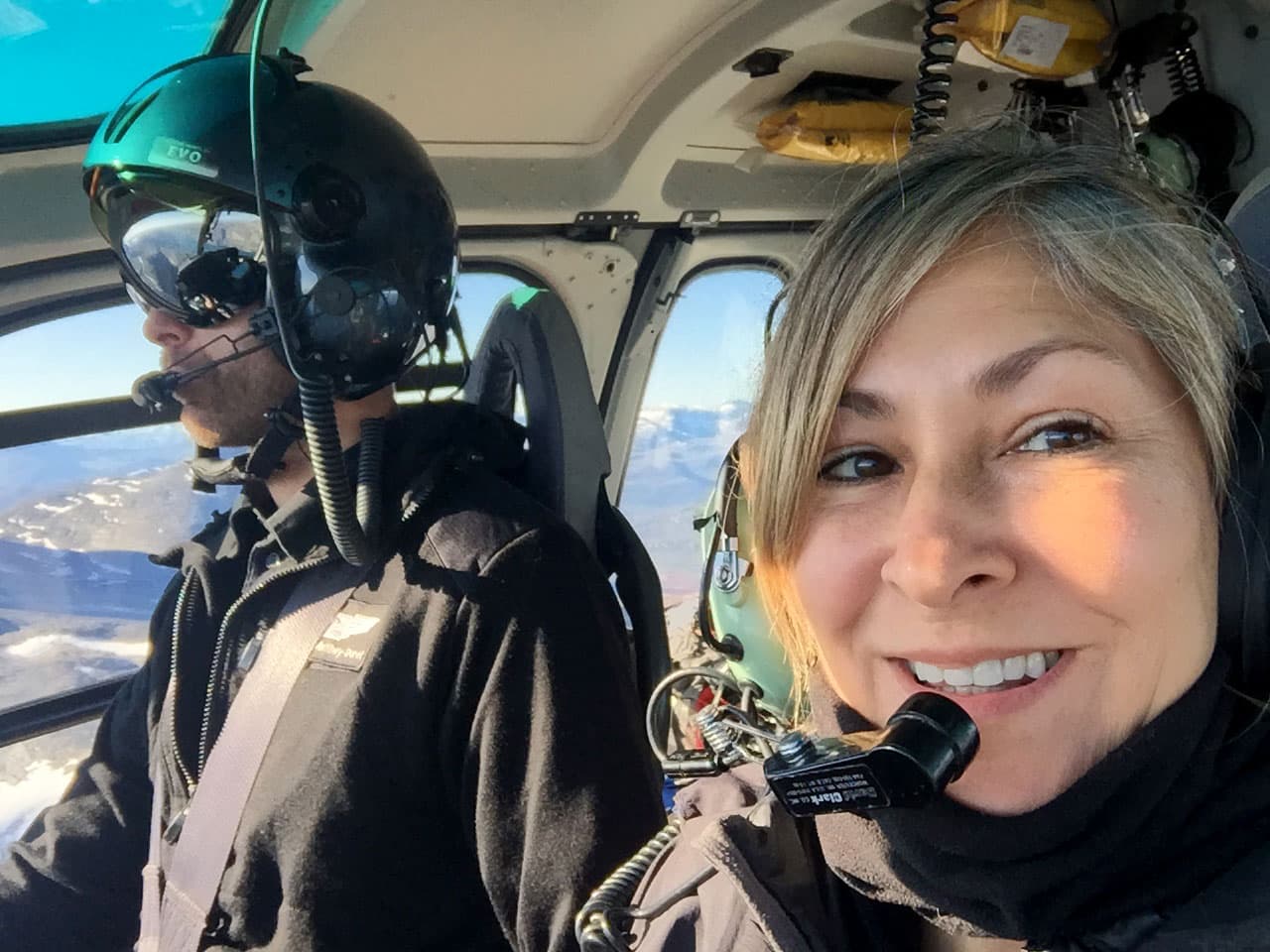 A woman wearing a headset smiles from inside a helicopter.