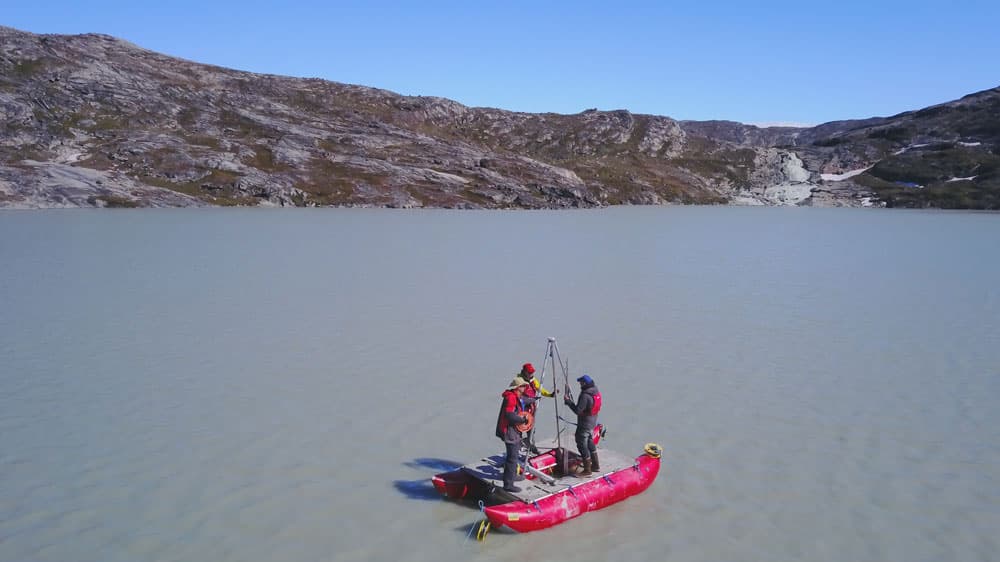 Three people stand on a bright red raft in the middle of a lake.