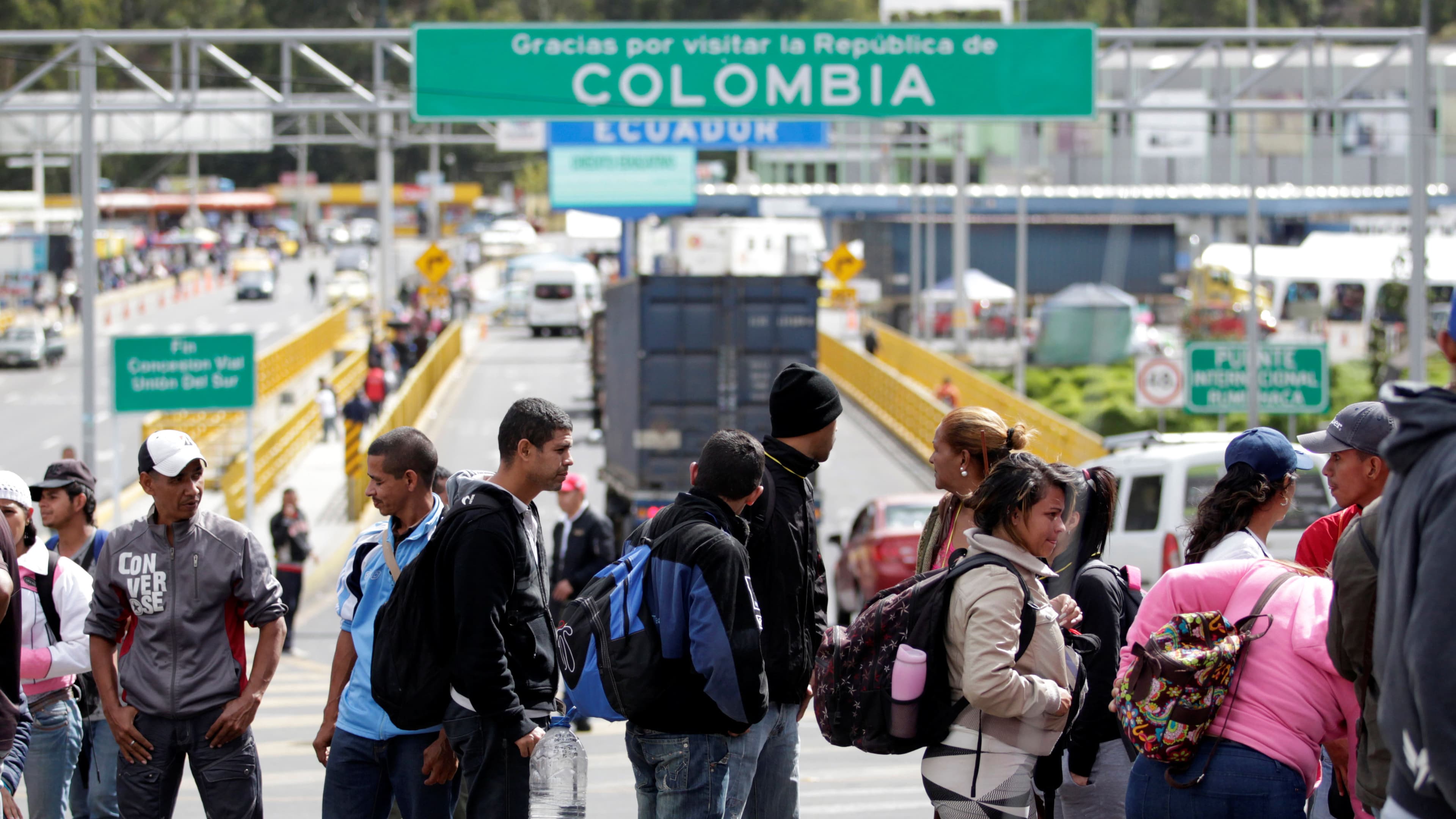 A line of migrants stand with backpacks and belongings under a green sign marking the Colombian border.