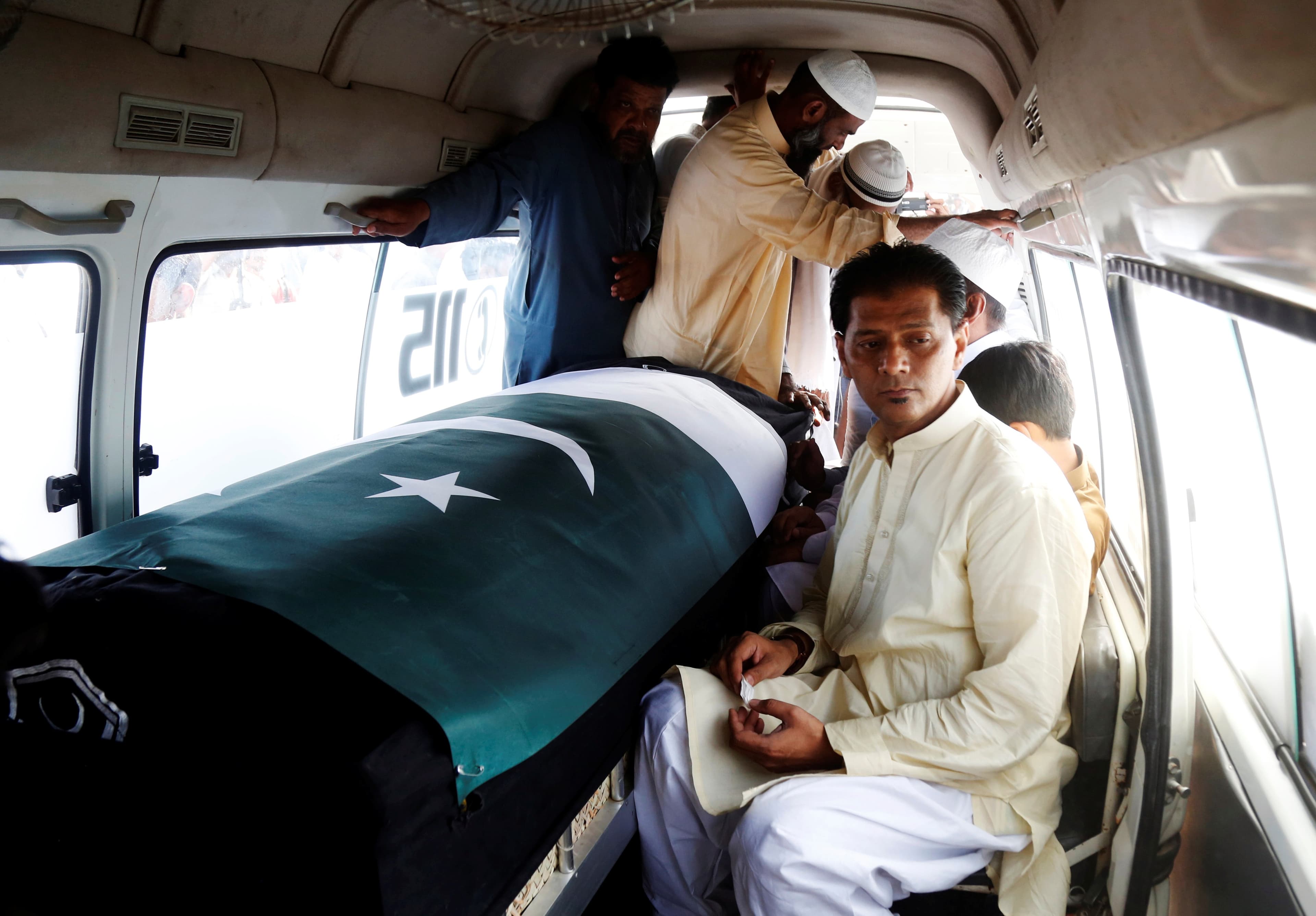 Man inside van sits next to coffin with Pakistan flag draped over, others standing nearby in close quarters