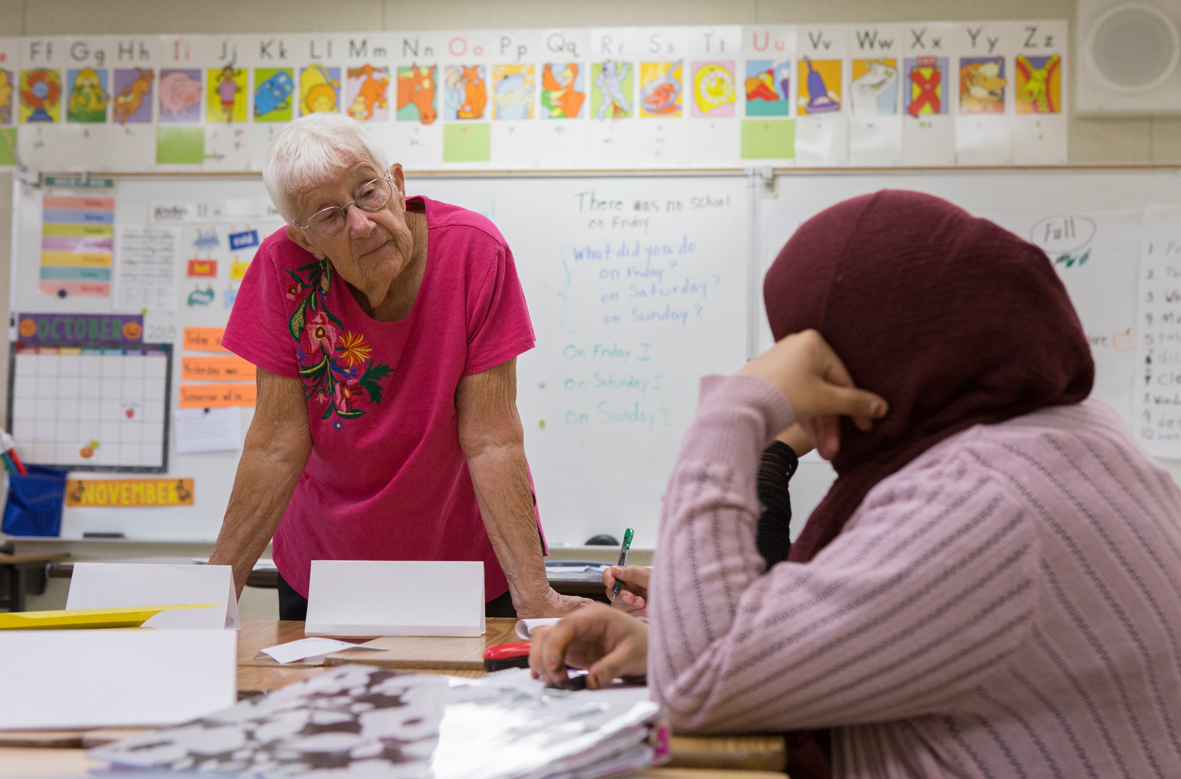 Instructor wearing a pink shirt, stands over a desk to speak to adult student who we see from behind