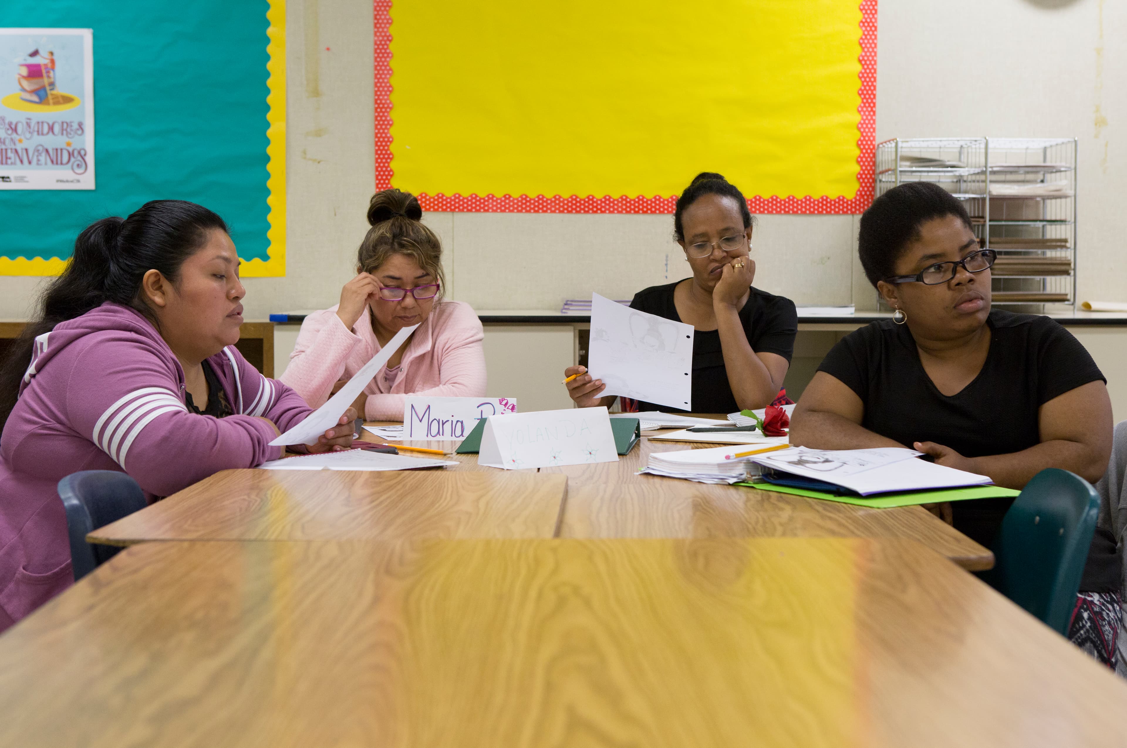 For women sit around elementary classroom tables, holding and reading papers