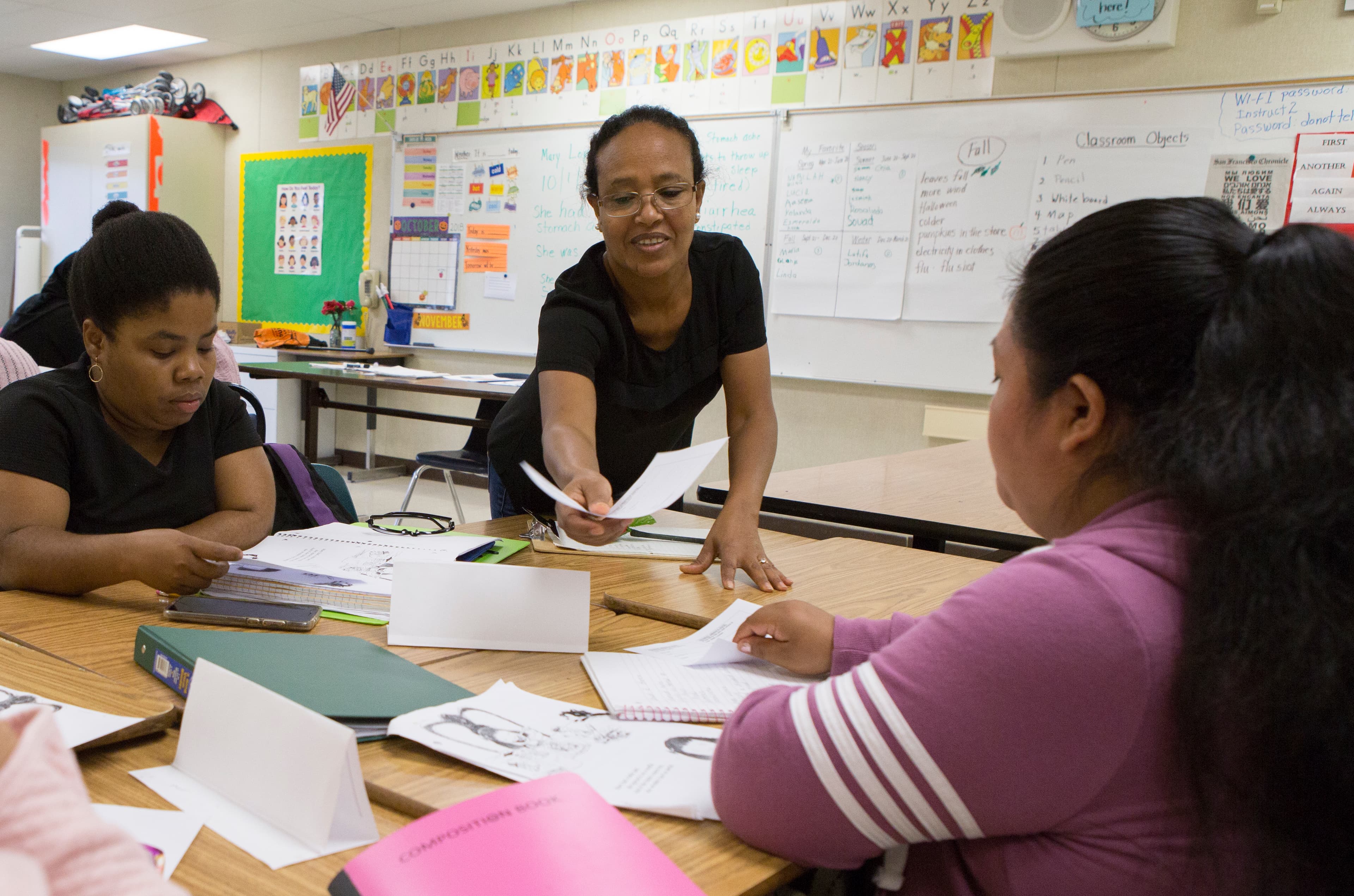 Woman leans over desks handing out paper, with two other women sitting around and white board in elemenatary school classroom behind them