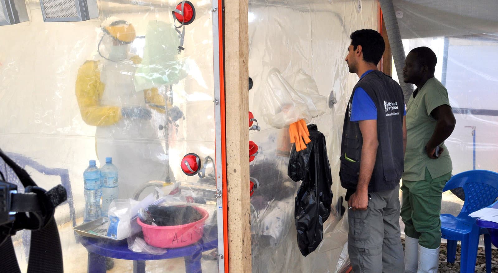 A doctor cares for a patient wearing protective clothing inside an isolate cube at a treatment center