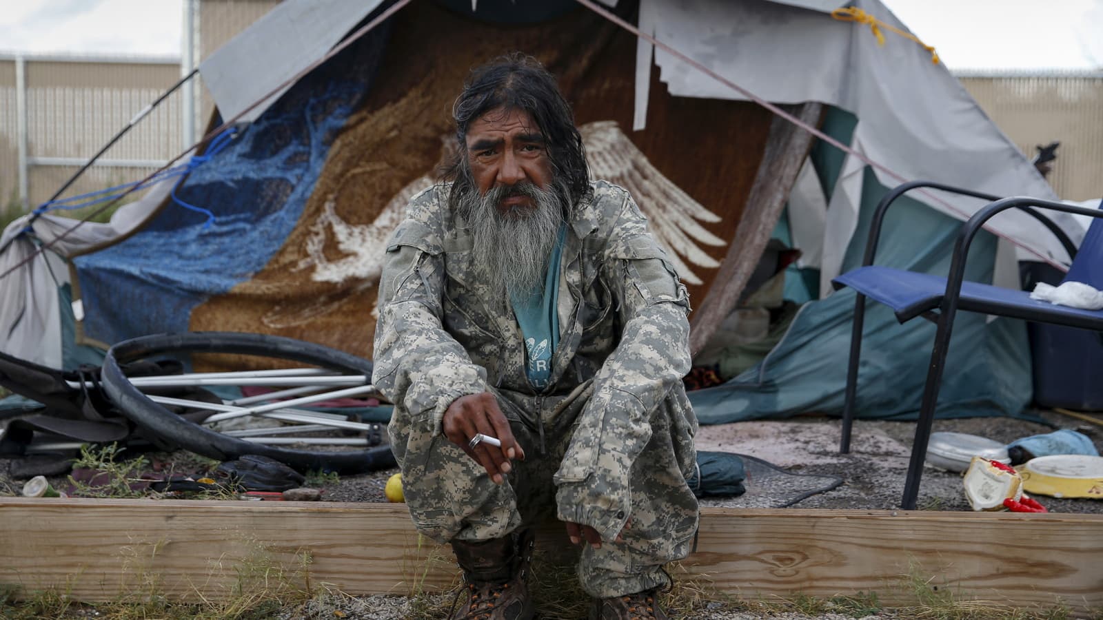 A bearded veteran sits outside a tent smoking a cigarette.